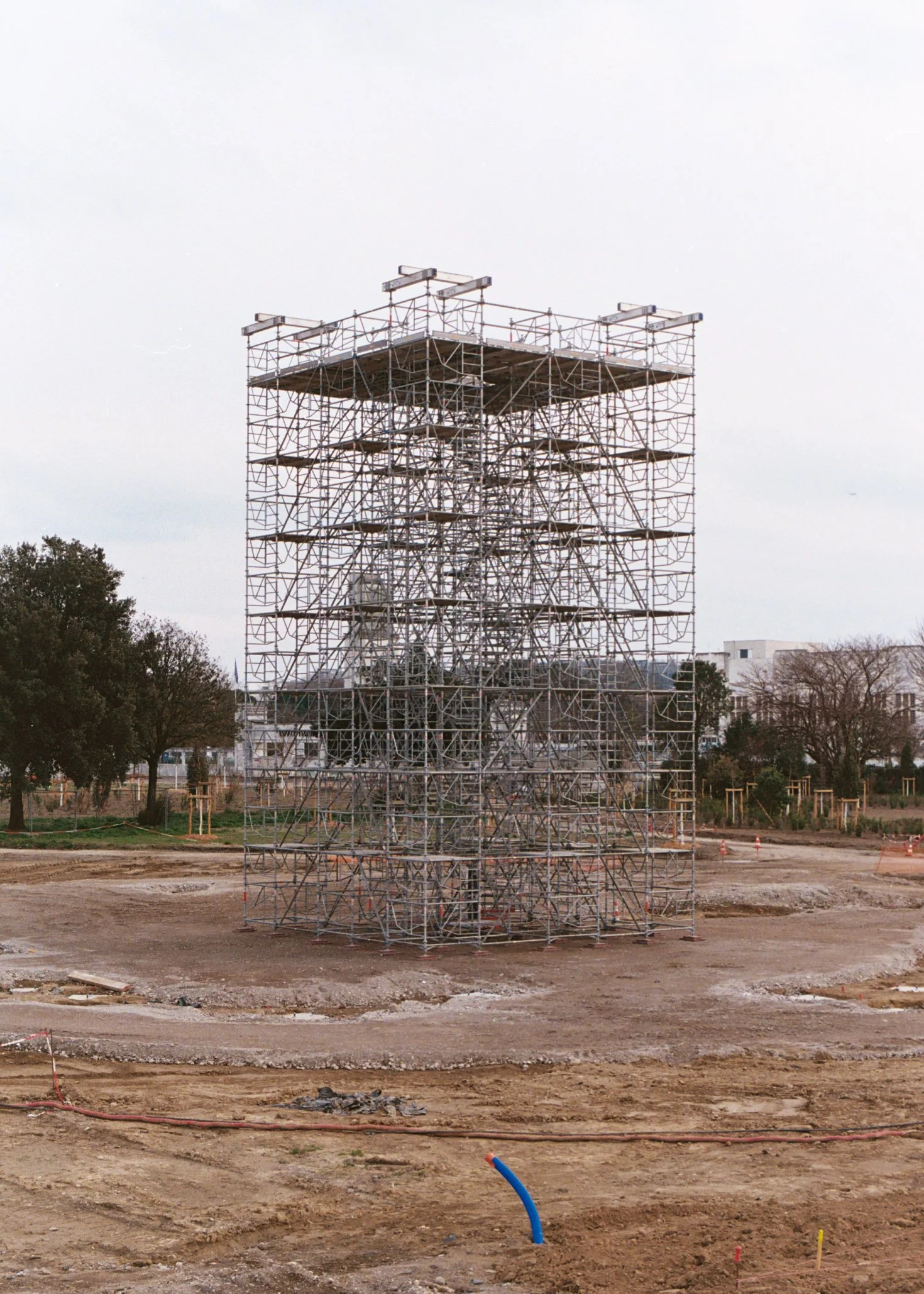 Une structure en métal de grande taille en cours de construction sur un terrain vide, avec des arbres et des bâtiments en arrière-plan.