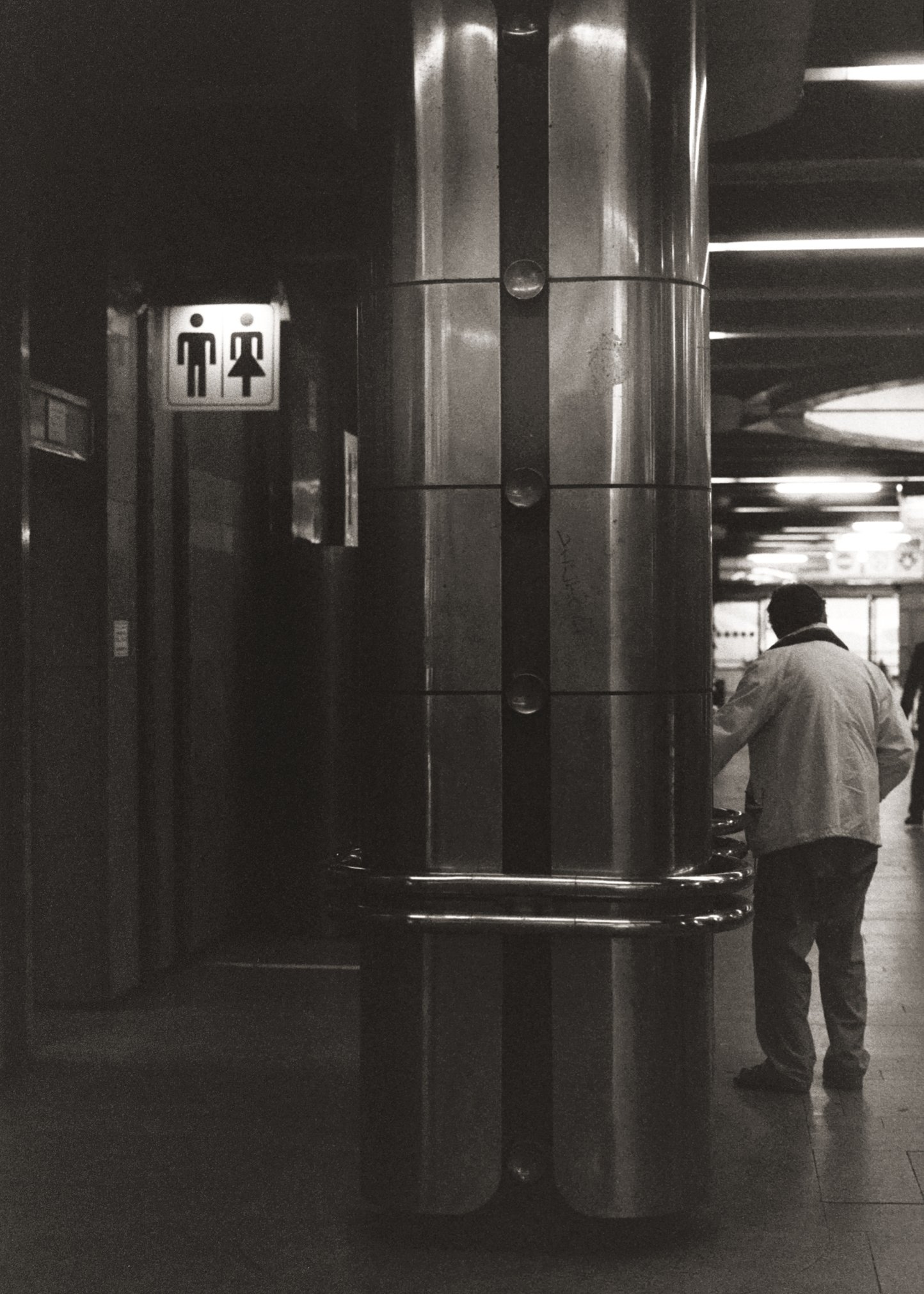 Une image en noir et blanc d'une sculpture rectangulaire en métal dans une station de métro. Deux personnes, un homme avec un manteau blanc et une autre personne vêtue de noir, se tiennent à proximité de la sculpture.