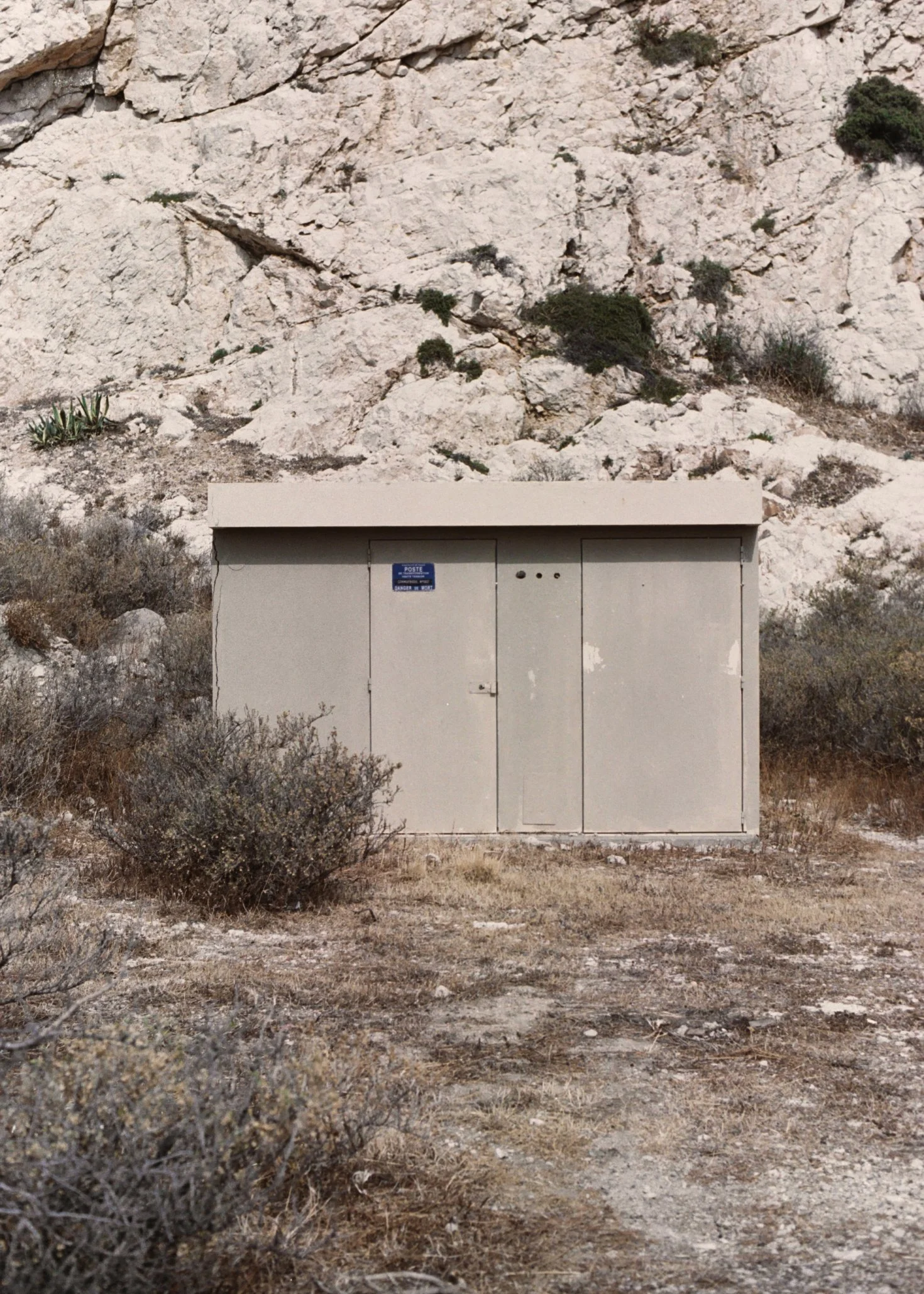Un bâtiment gris métallique, semblable à une boîte aux lettres ou un boîtier électrique, situé devant une roche calcaire aride avec des buissons et des plantes désertiques.