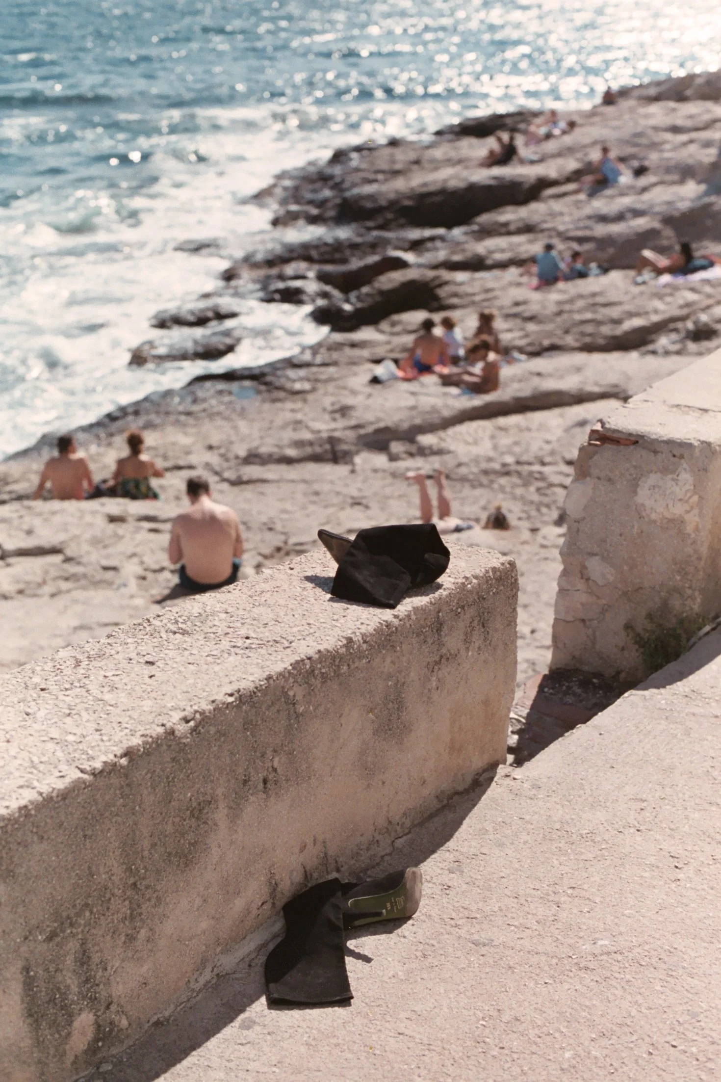 Vue d'une plage rocheuse avec des personnes qui se détendent au soleil, vue partielle d'une chaussure noire, un sac et un objet sur un mur en pierre en premier plan.