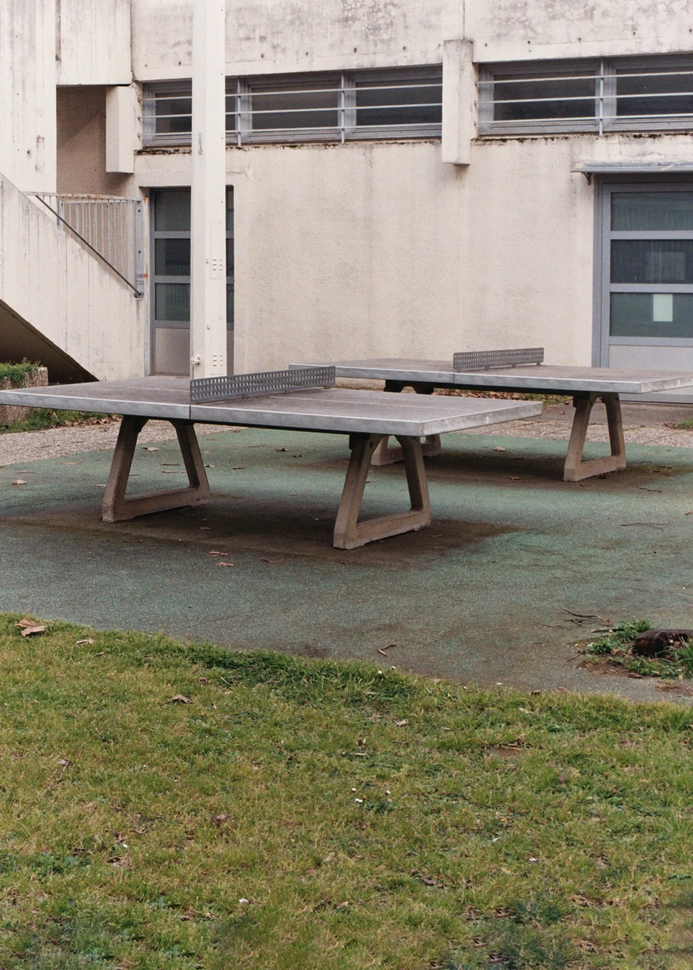 Deux tables de ping-pong en béton dans une cour extérieure, entourées de pelouse et d'un bâtiment moderne en arrière-plan.