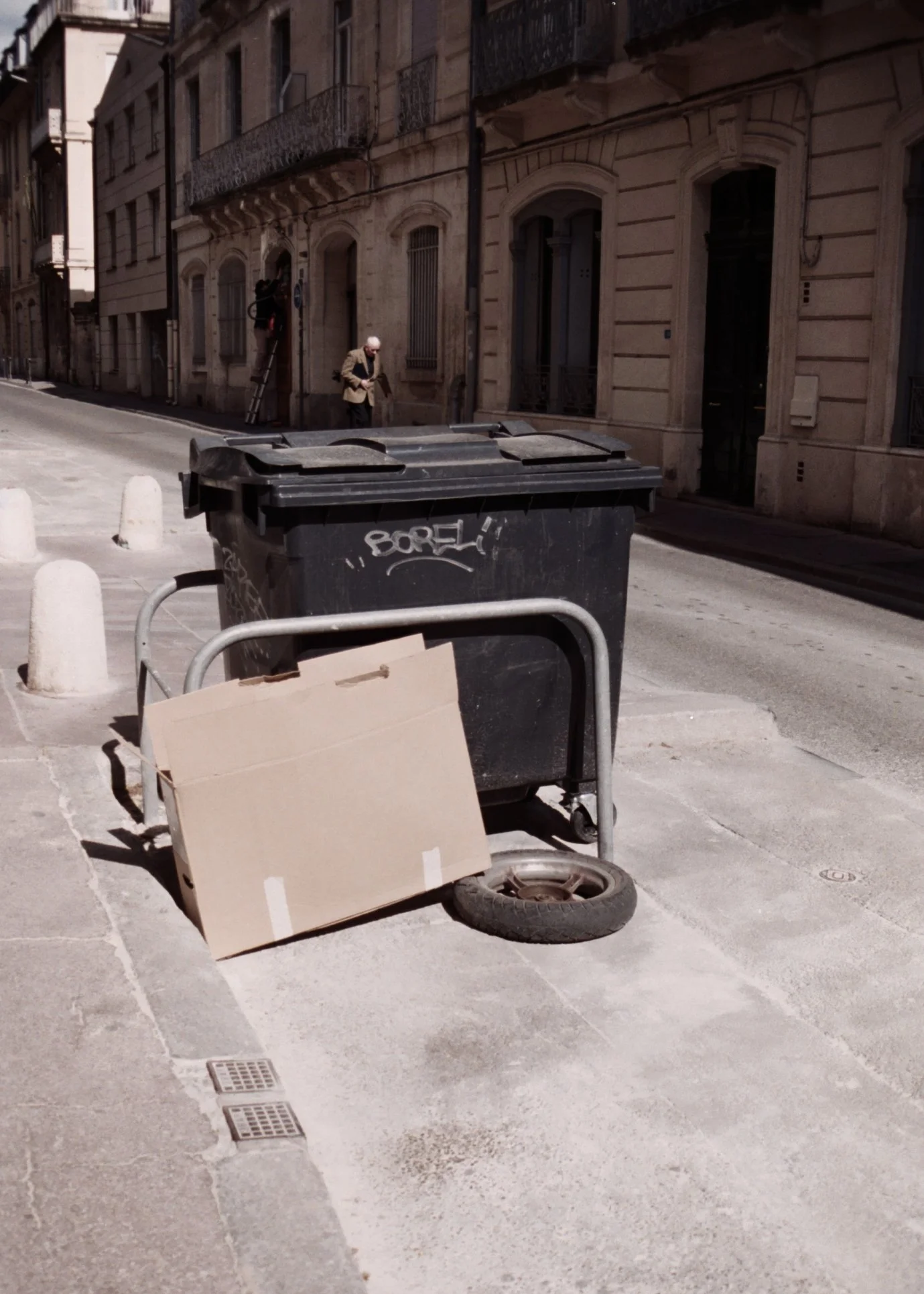 Une poubelle noire avec graffiti, un chariot à déchets avec une grande boîte en carton, un pneu usé, une chaussée en pierre, un mur en pierre d'un bâtiment ancien, un homme âgé portant un manteau beige marchant dans une rue urbaine, des barrières en 