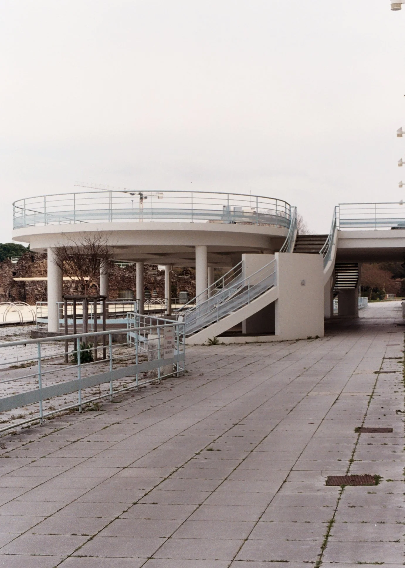 Un bâtiment moderne avec une plateforme ronde surélevée accessible par un escalier extérieur. La scène est vide, avec un sol en pavés et quelques arbres en arrière-plan.