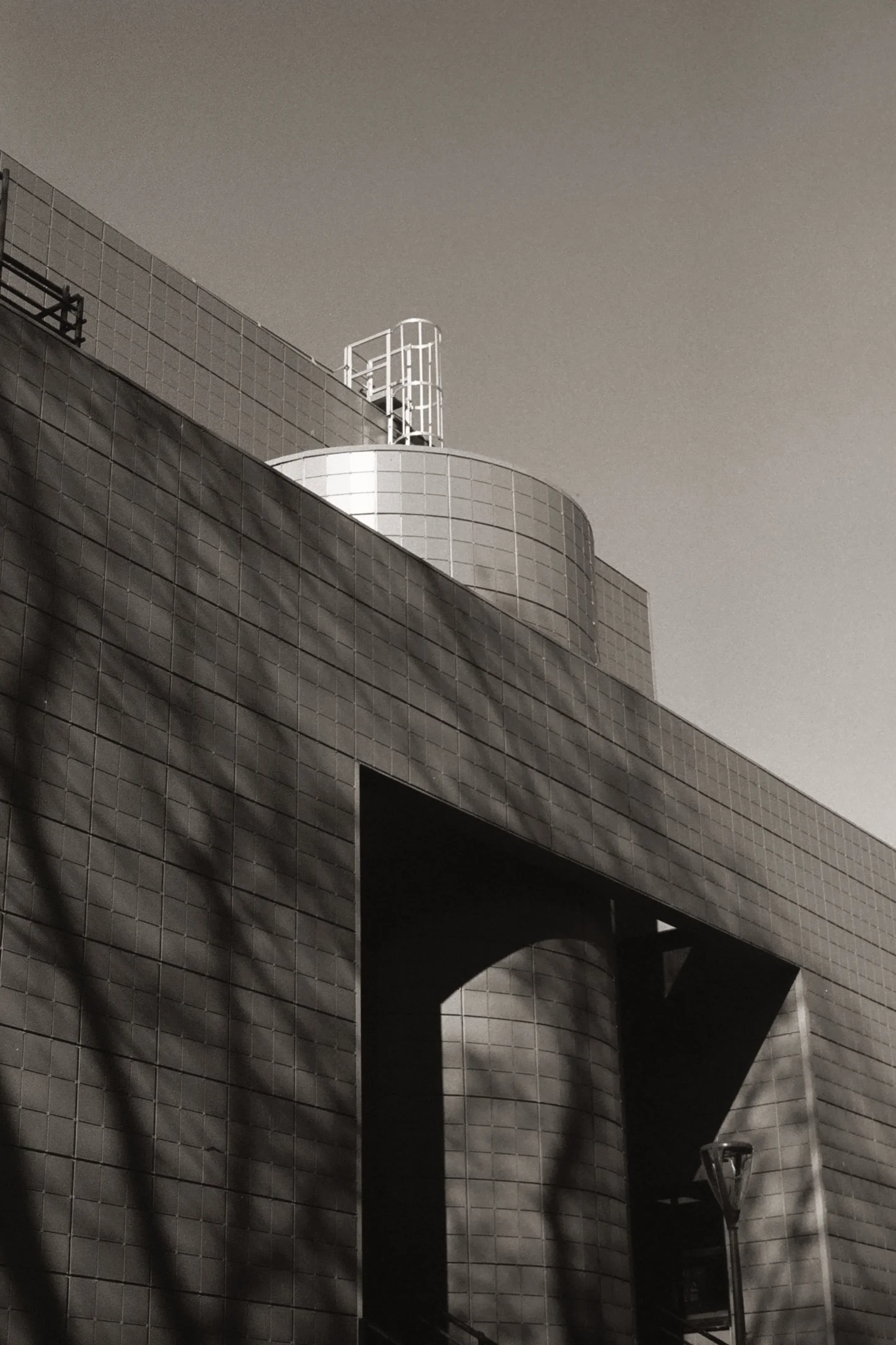 Une photographie en noir et blanc d'un bâtiment moderne en hauteur avec une façade en carreaux et une terrasse en haut. Un portail en fer blanc au sommet de la structure. Un lampadaire se trouve dans la zone inférieure de l'image.