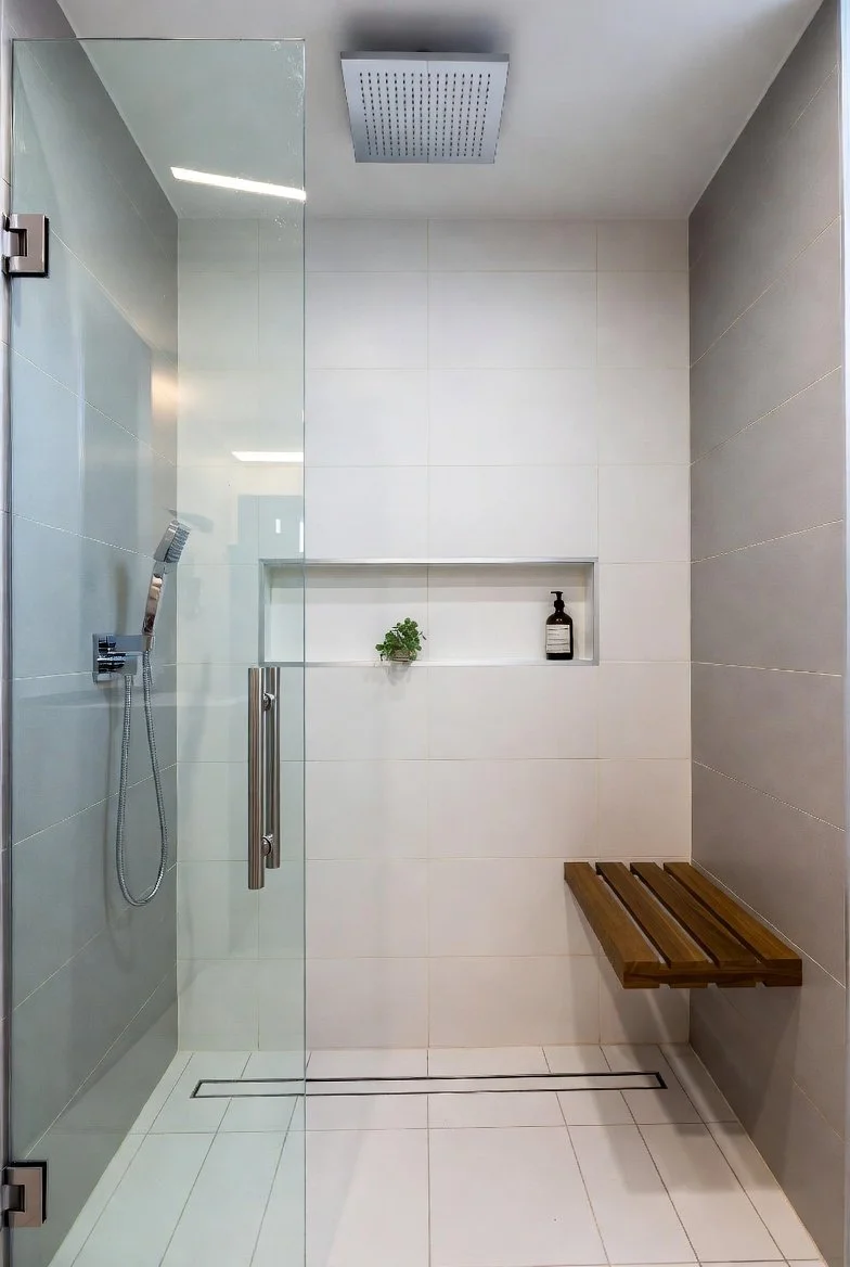 A modern walk-in shower with a glass door, a rain showerhead on the ceiling, a wall-mounted handheld showerhead, a built-in shelf with a small plant and a bottle of soap, a wooden bench, and a linear drain on the floor.