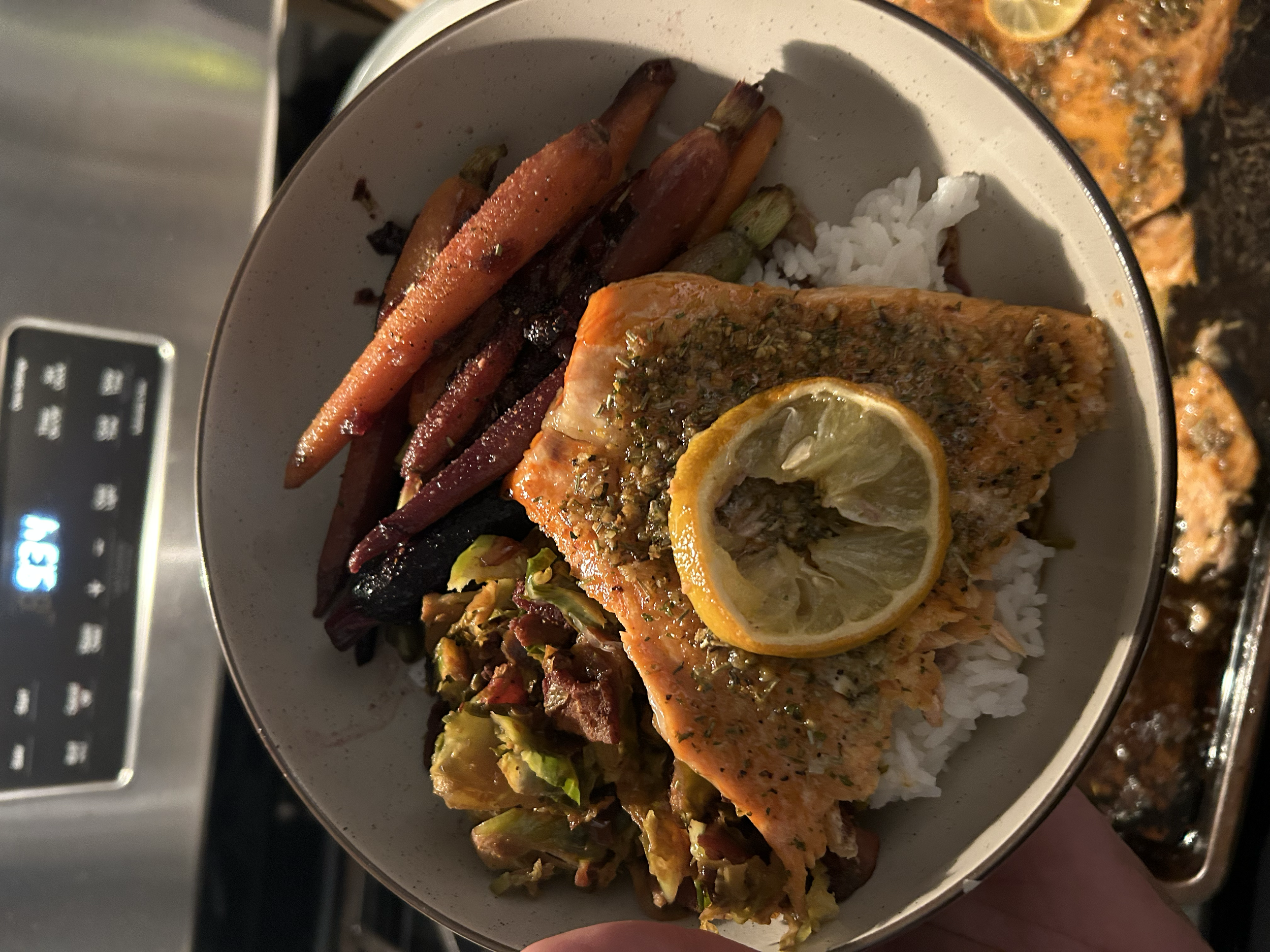 A bowl of cooked salmon topped with lemon and herbs, served over white rice with roasted vegetables on the side, including carrots and eggplant.