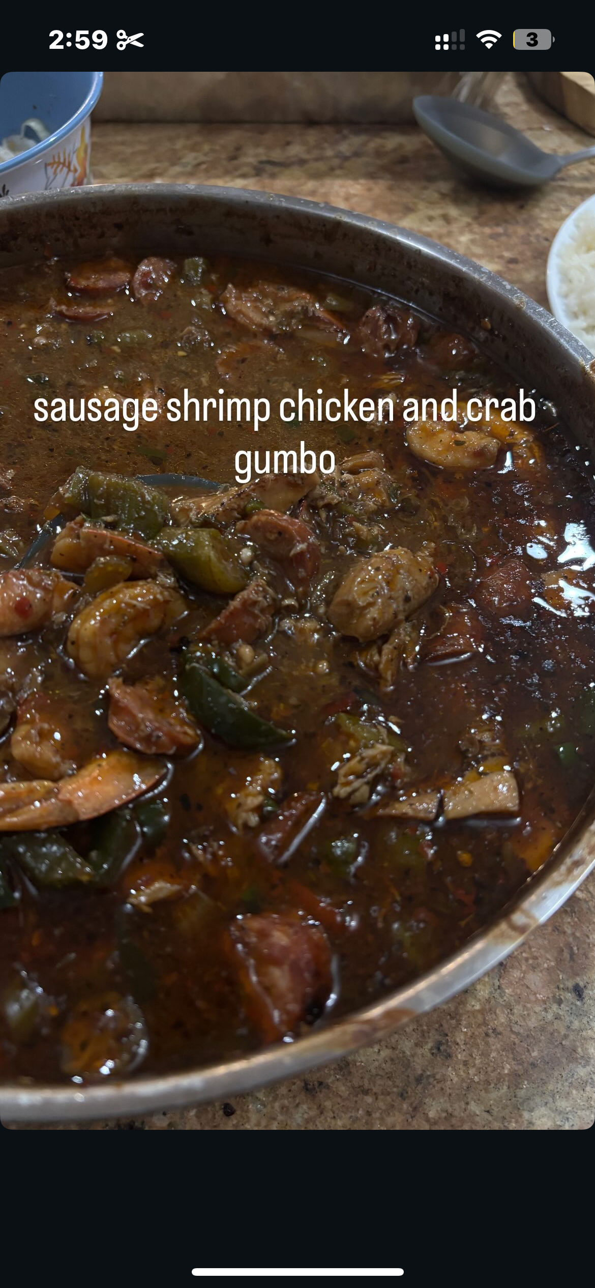 A bowl of sausage, shrimp, chicken, crab, and gumbo with visible vegetables and spices on a wooden table.