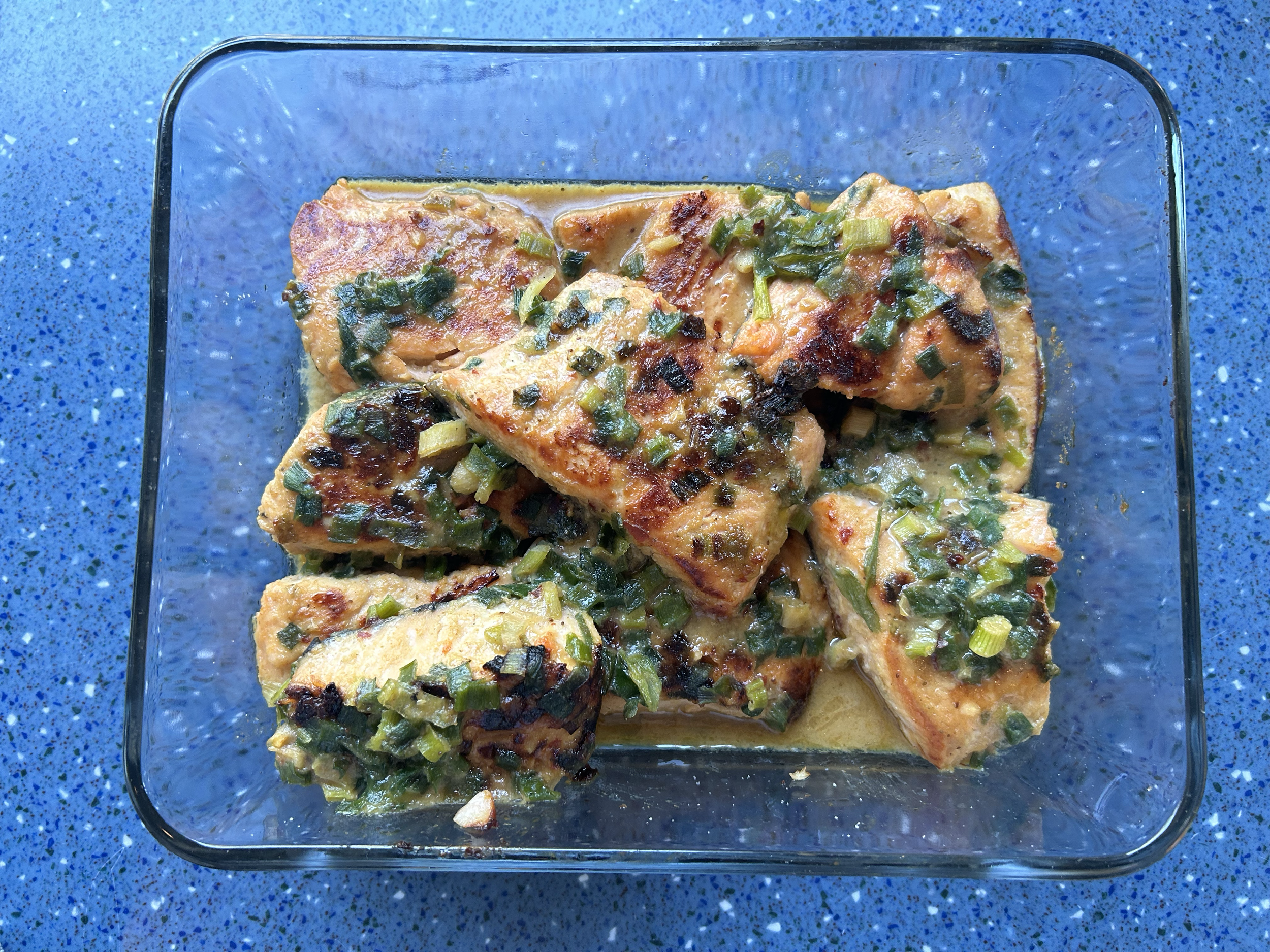 Pan-fried fish fillets in a creamy sauce with chopped green onions in a glass dish on a blue speckled countertop.