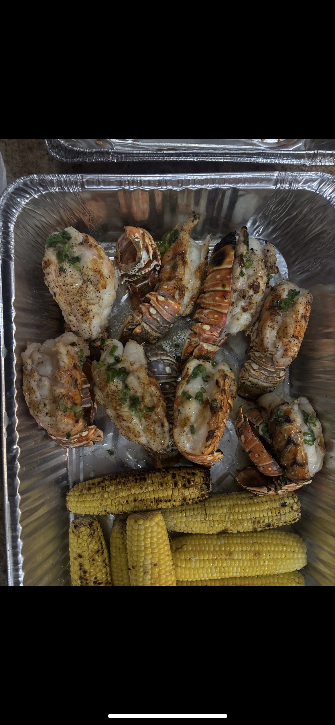 Aluminum tray containing grilled lobster tails garnished with green onions and grilled corn on the cob.
