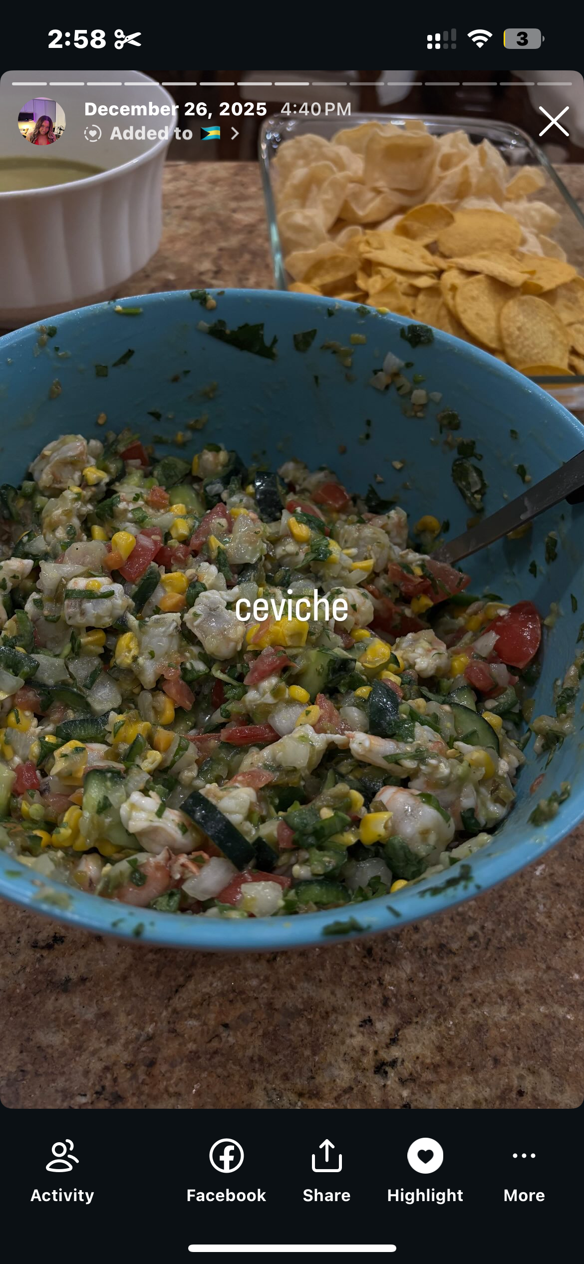 A bowl of ceviche with chopped vegetables, corn, and seafood on a kitchen counter, with potato chips in a glass dish and a cup of green sauce in the background.