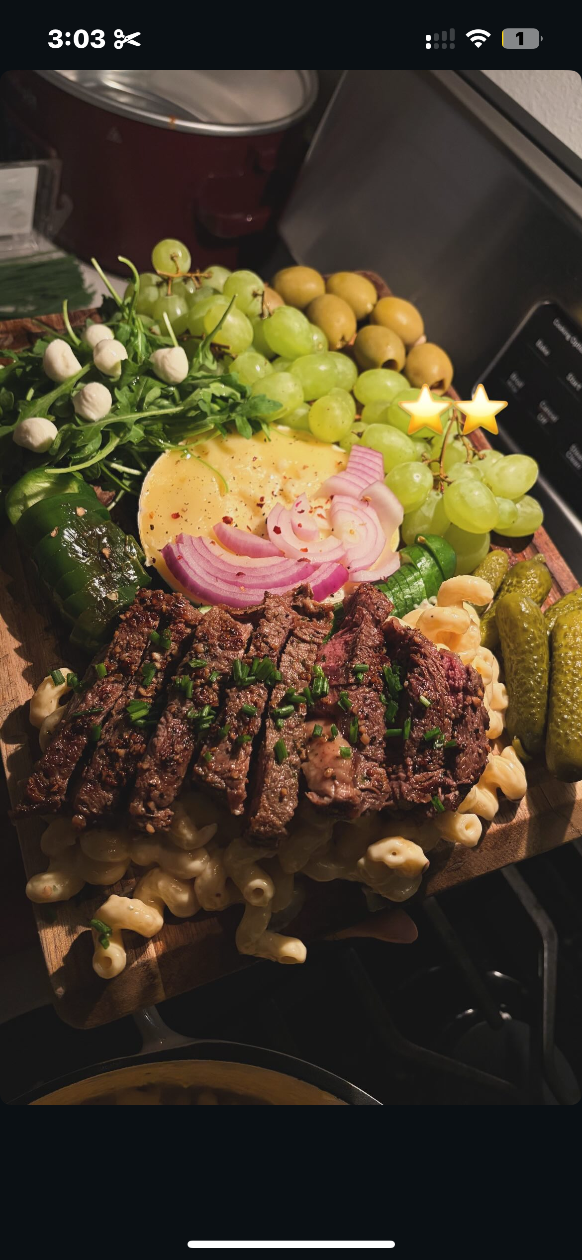 A wooden serving board with sliced steak, macaroni salad, grapes, cheese, onions, pickles, and greens.