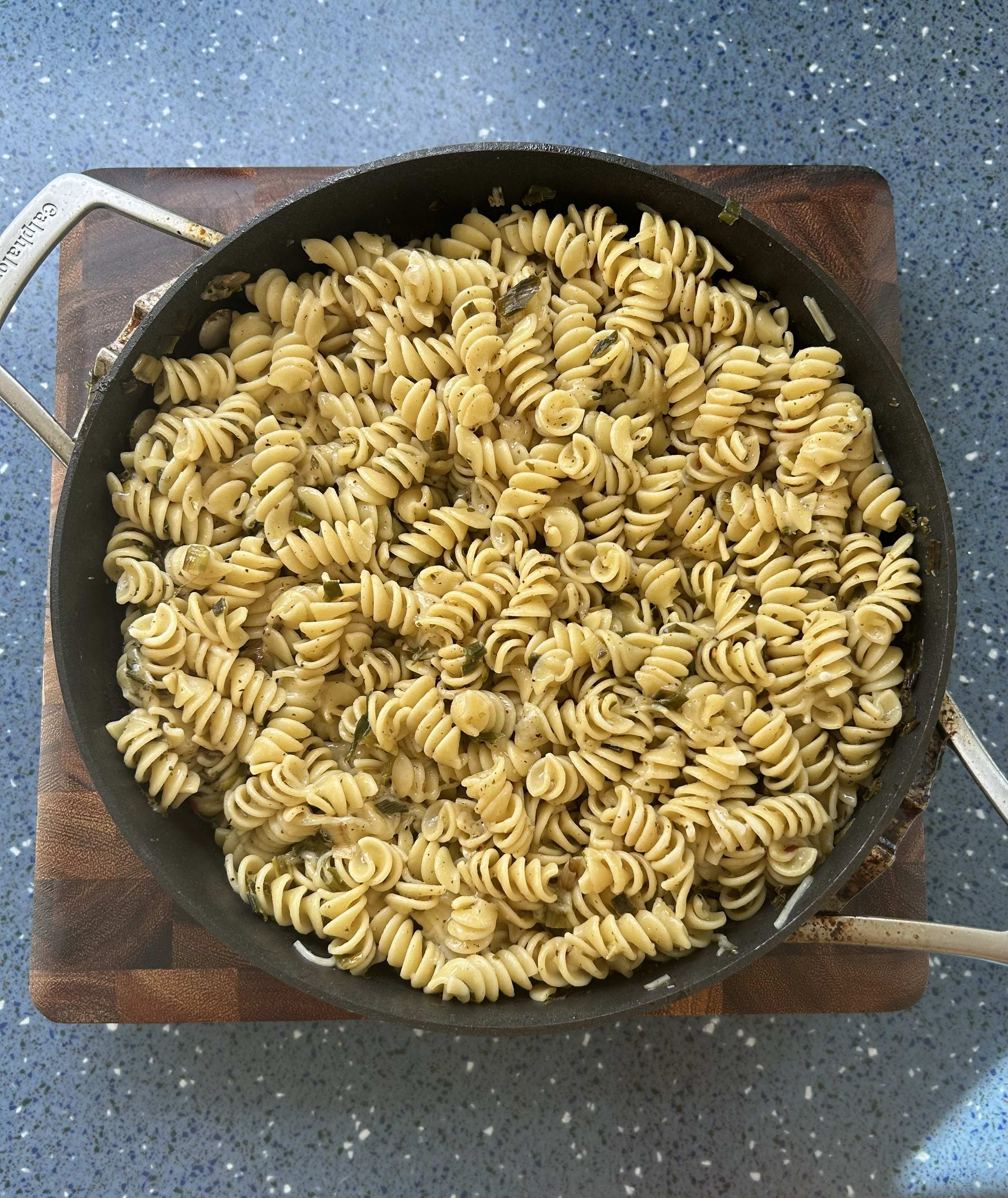 Fusilli pasta with herbs in a black skillet on a wooden cutting board, resting on a speckled blue countertop.