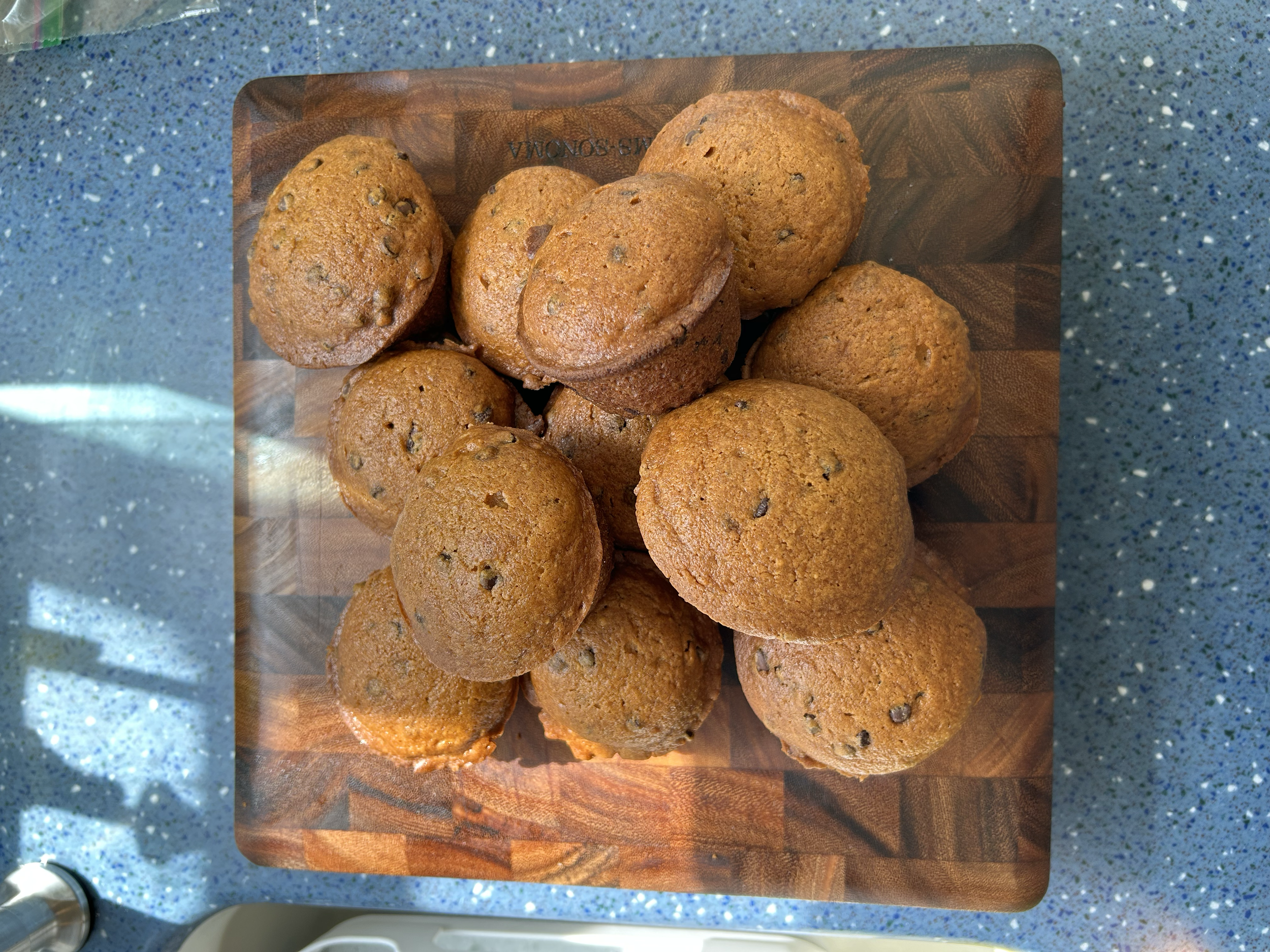 A wooden cutting board with twelve chocolate chip muffins on a blue speckled countertop.