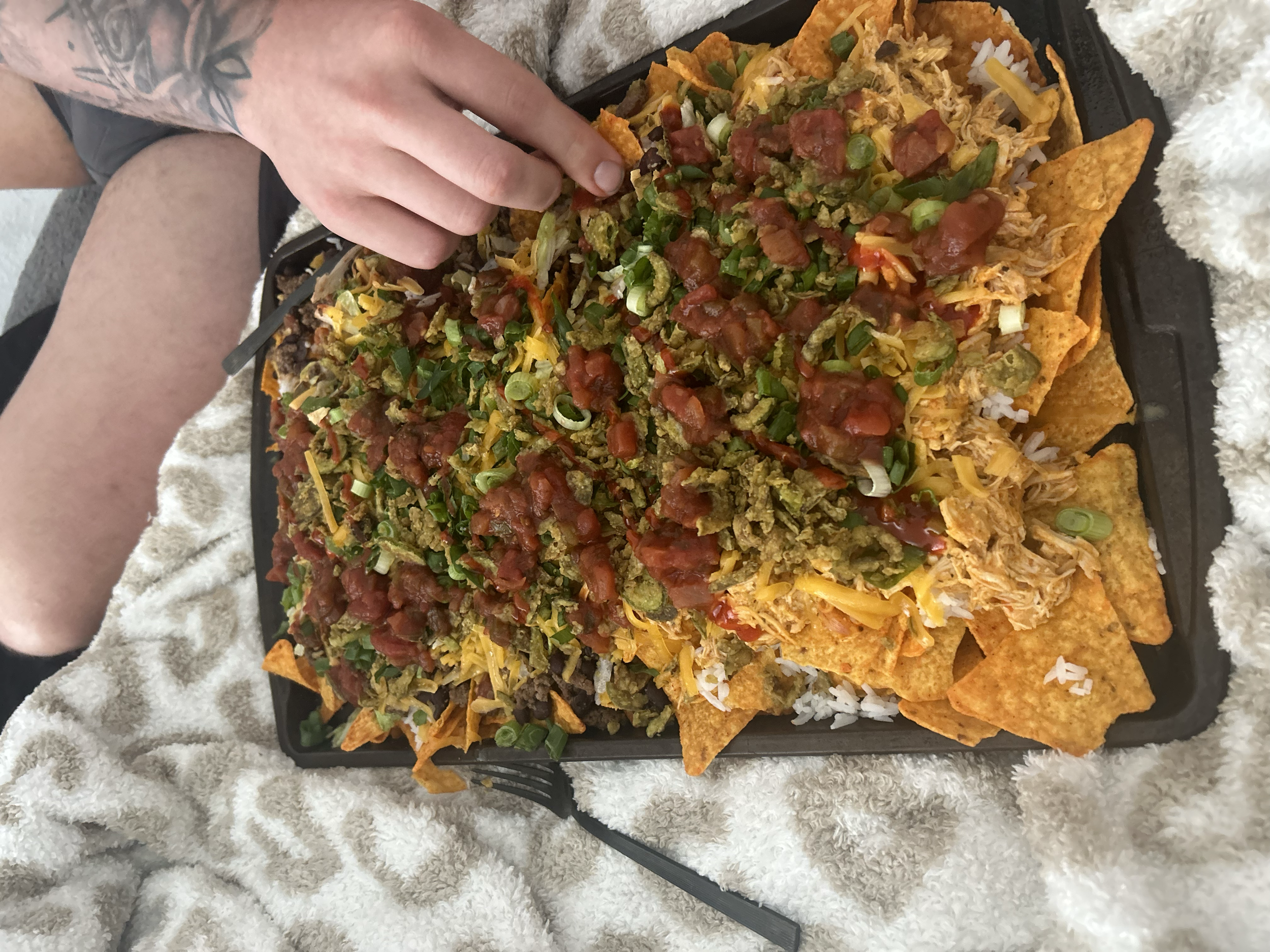 A tray of nachos with cheese, shredded chicken, ground beef, guacamole, diced tomatoes, chopped green onions, and salsa, with a person's hand reaching to add toppings.