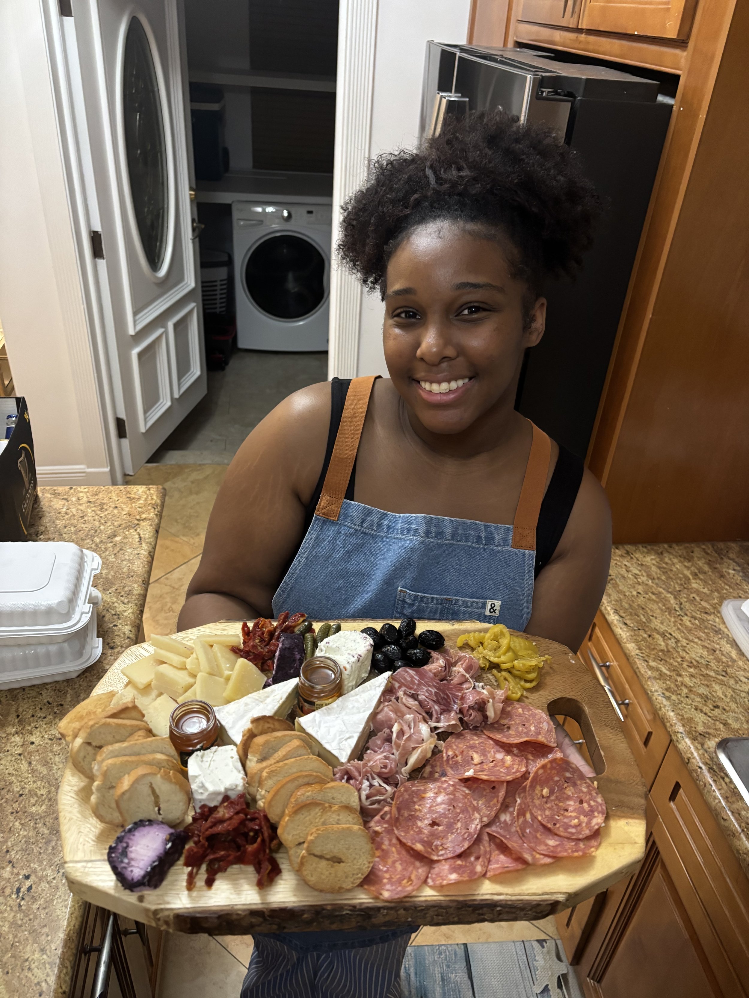 A woman smiling and holding a large cheese and charcuterie board with various cheeses, meats, crackers, olives, and condiments in a kitchen.