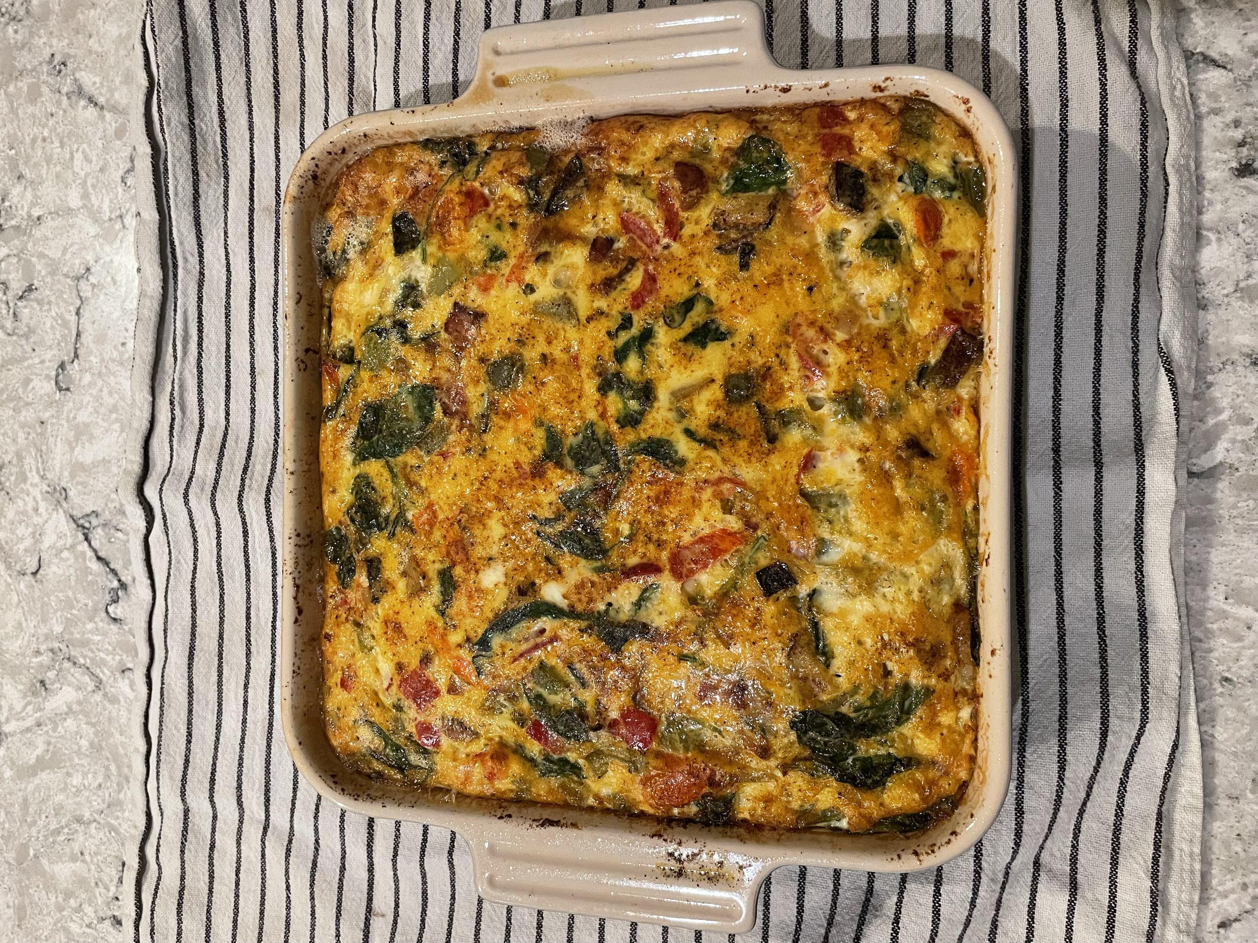 Baked casserole with vegetables and cheese in a square dish on a striped cloth.