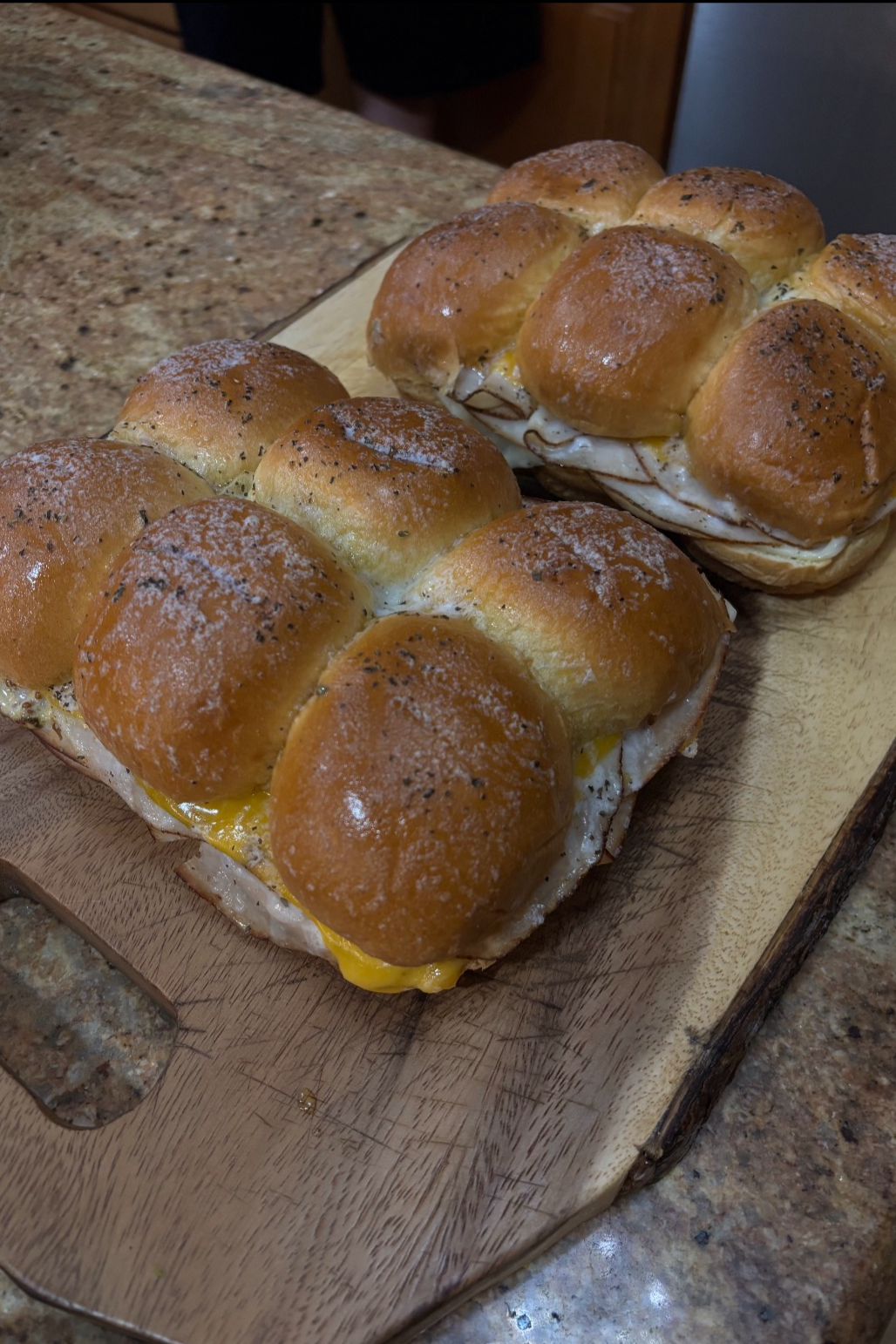 Two large breakfast sandwiches on a wooden cutting board, each with soft buns, melted cheese, and egg. The sandwiches are sprinkled with black pepper and salt.