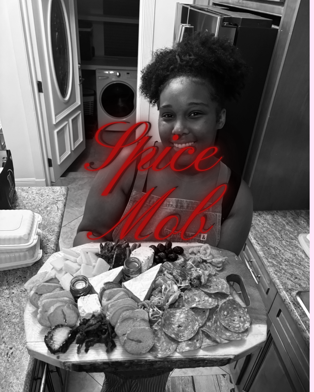 A woman with natural curly hair smiling at the camera, standing in a kitchen behind a large tray of assorted food including grilled vegetables, chips, sliced meats, and small jars, with a washing machine visible in the background. The photo has the w