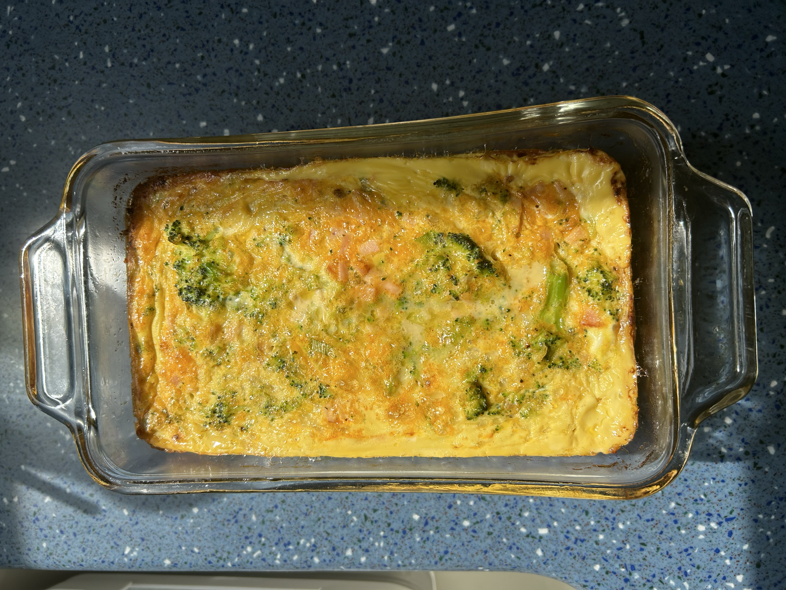 Baked casserole in a glass dish on a speckled blue countertop, with visible broccoli, cheese, and possibly other vegetables.