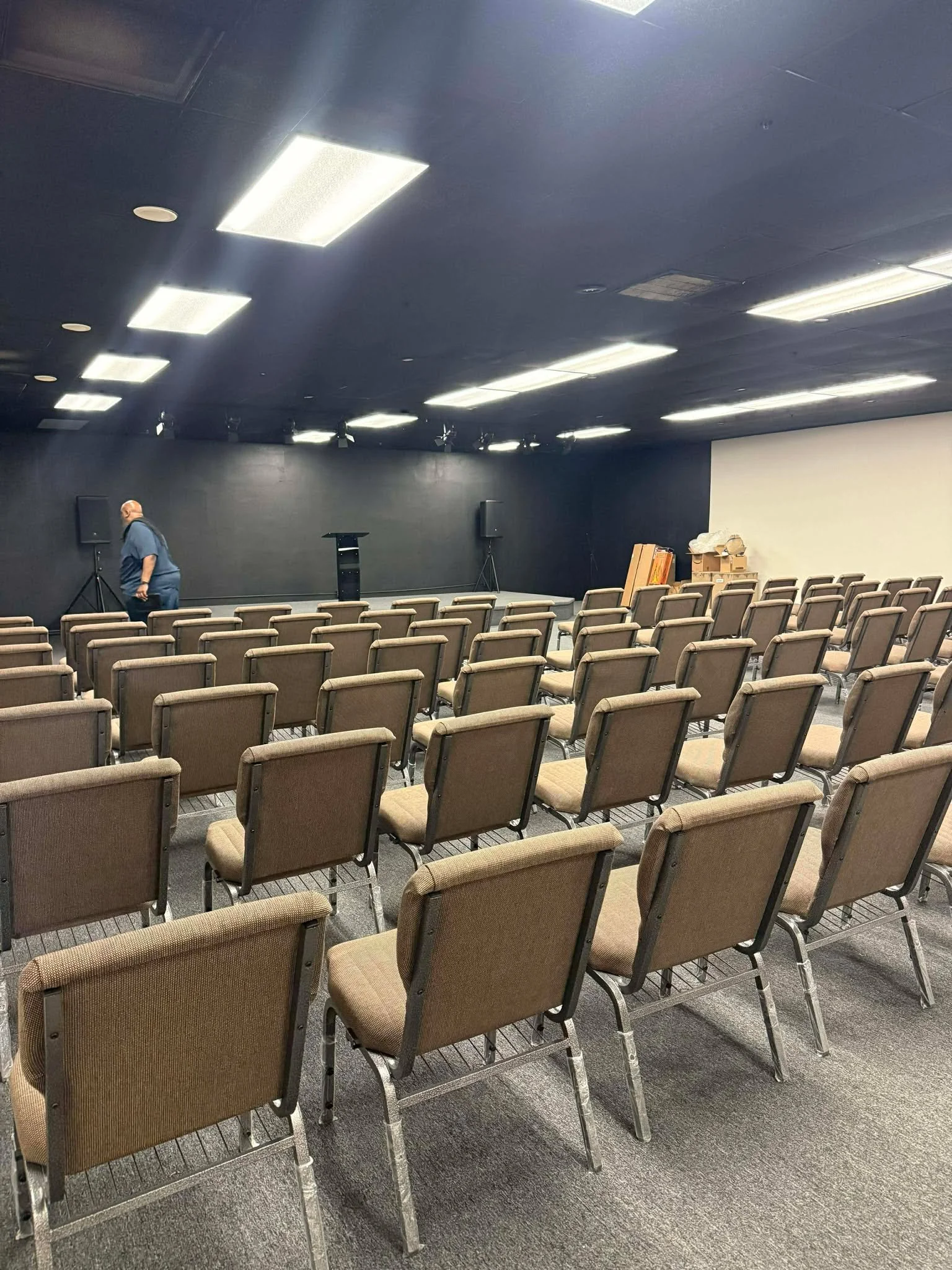Empty sanctuary with rows of beige chairs facing a stage with a man on the left side, black wall, and stacked items on the right.