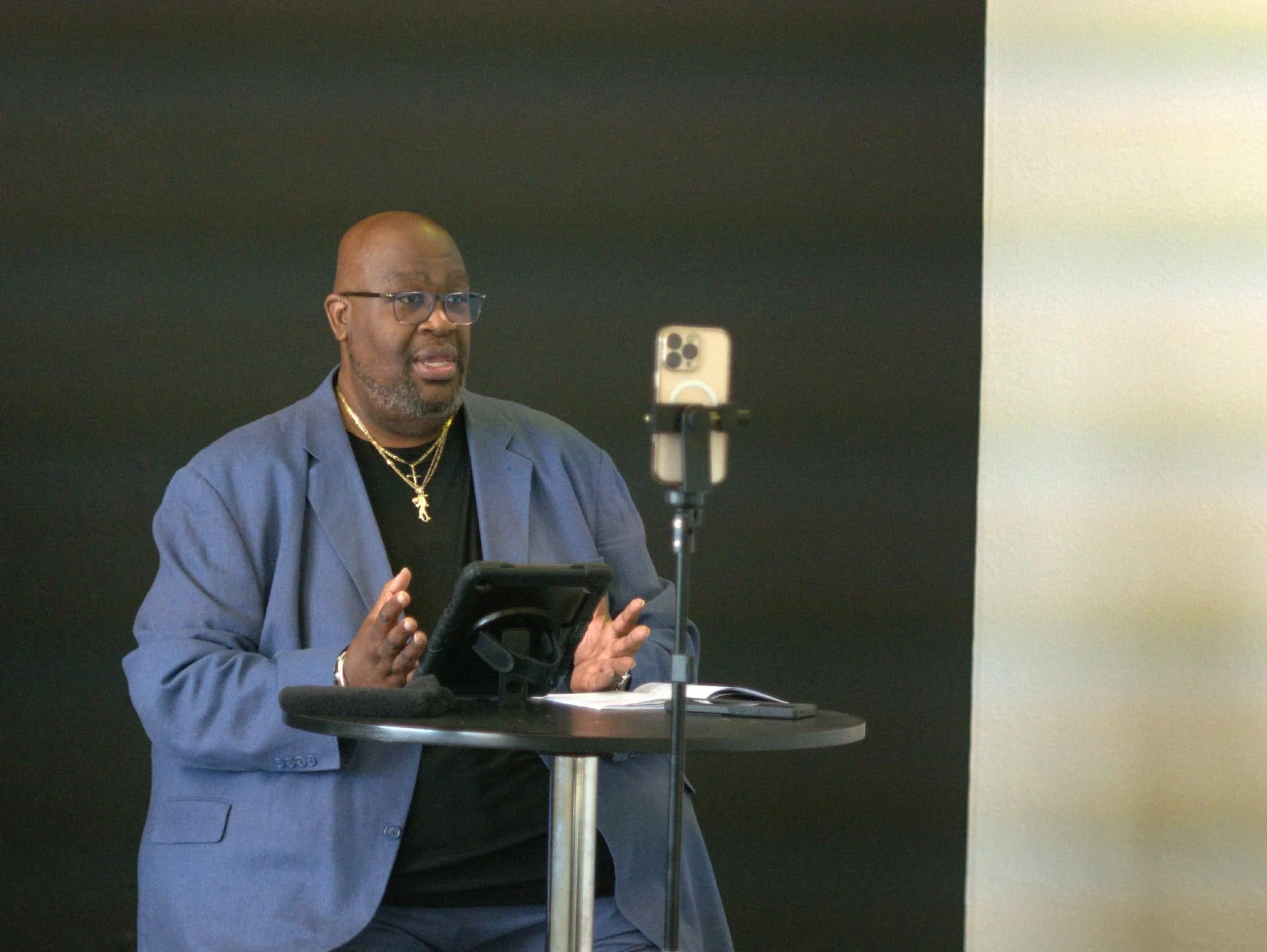A man sitting at a round table, speaking and gesturing with his hands, while looking at a smartphone mounted on a stand. He is wearing glasses, a blue blazer, a black shirt with gold necklaces, and is positioned against a dark background with a beige wall to his right.