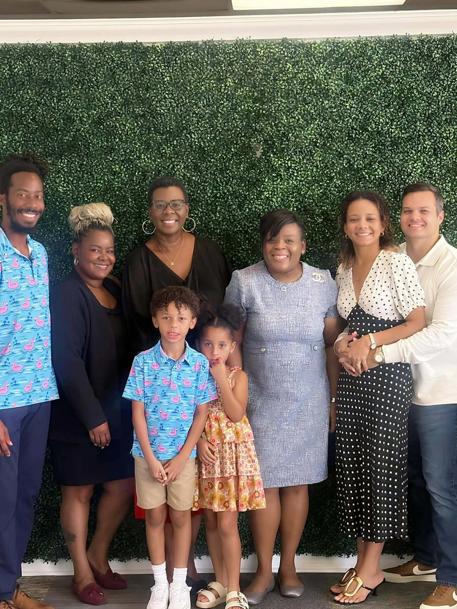 A diverse group of nine people, including three children and six adults, standing together indoors in front of a green leafy wall, smiling for a group photo.