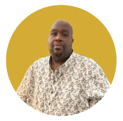 Portrait of a man with a shaved head and beard, wearing a floral button-up shirt, against a background with a yellow circle and light blue outer area.