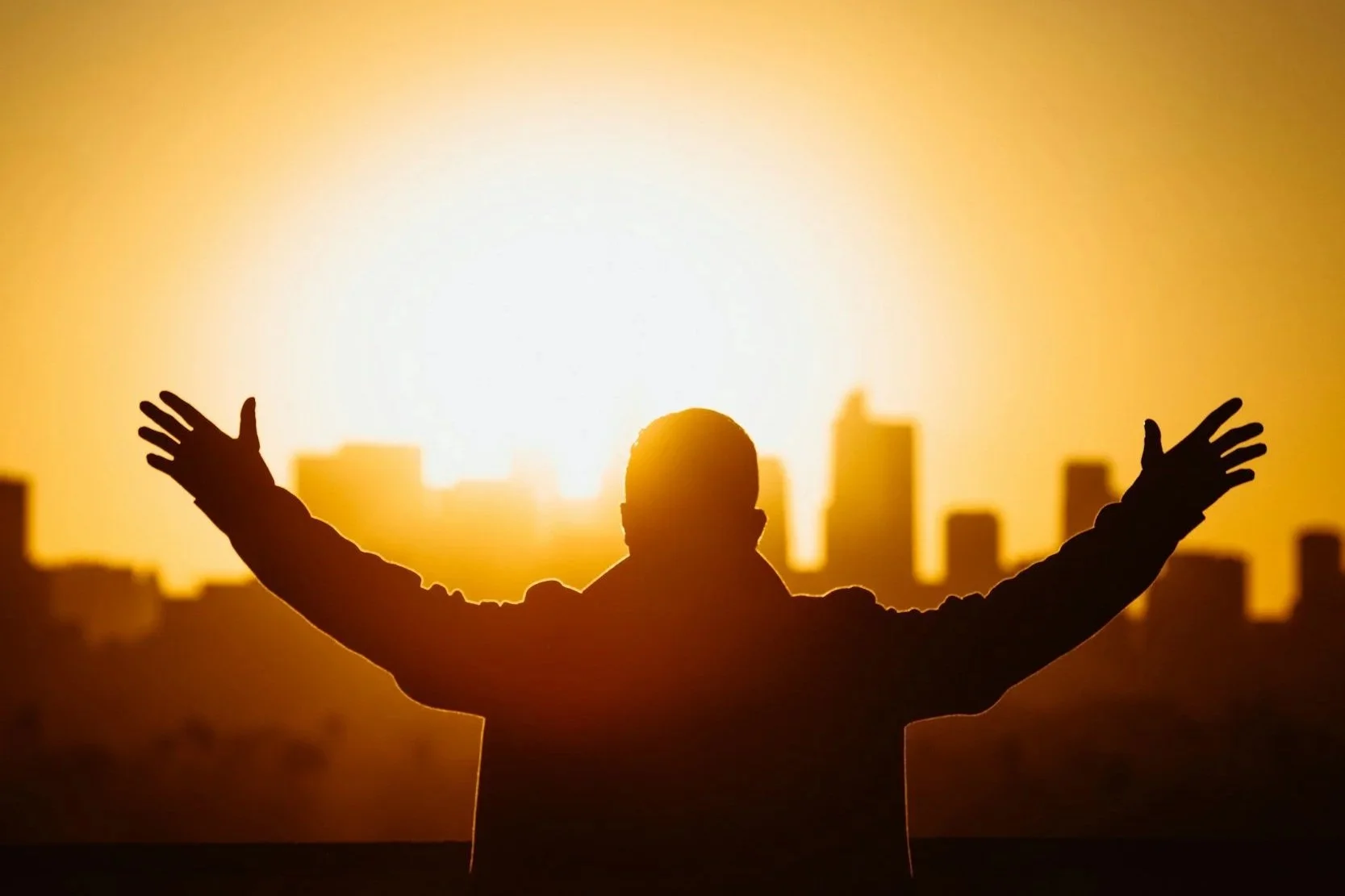 Silhouette of a person with arms outstretched standing against a bright sunset with city buildings in the background.