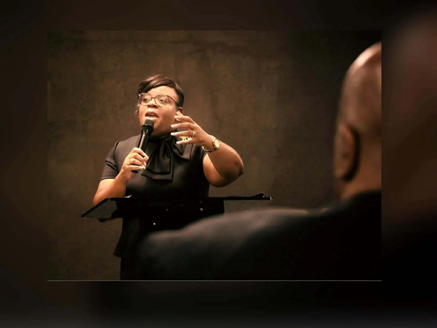 A woman speaking into a microphone at a podium, gesturing with her hand during a presentation or speech.