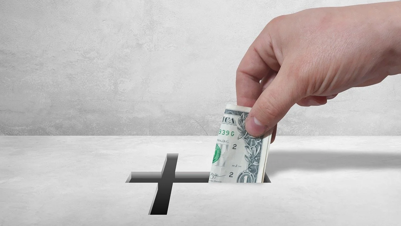 A hand placing a dollar bill into a hole with a cross cutout, resembling a donation or contribution.