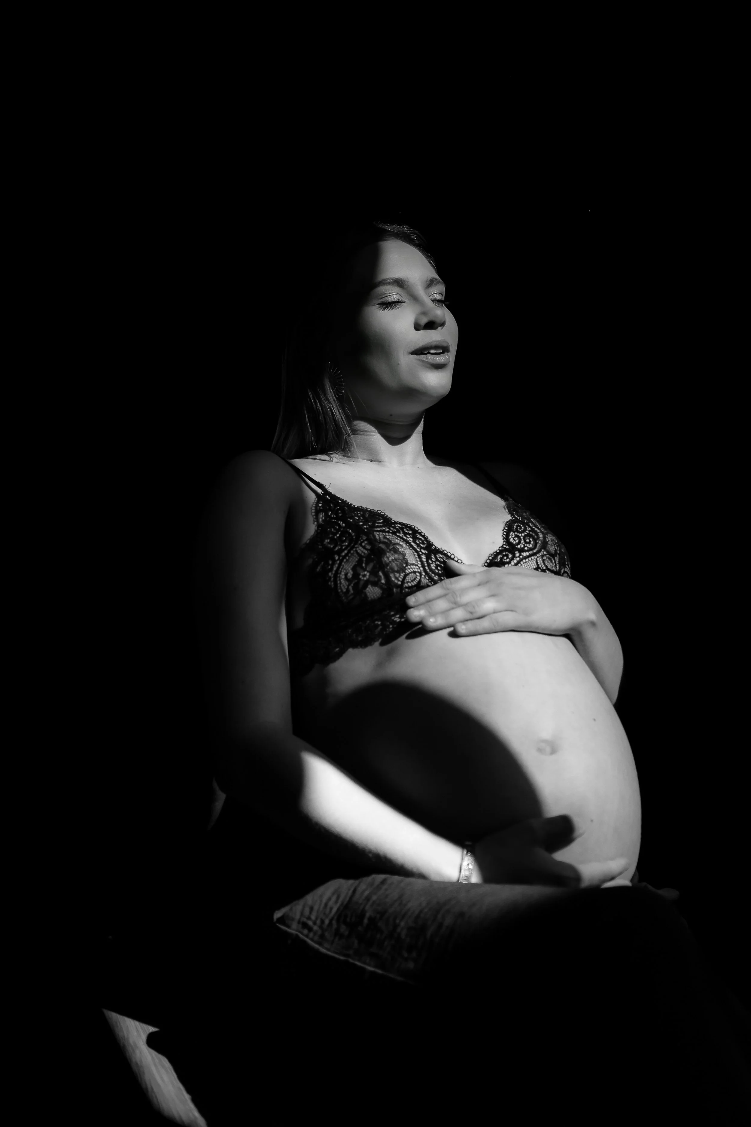 Femme enceinte habillée en lingerie en dentelle, assise avec une main sur le ventre, silhouette sombre en noir et blanc, expression de sérénité.