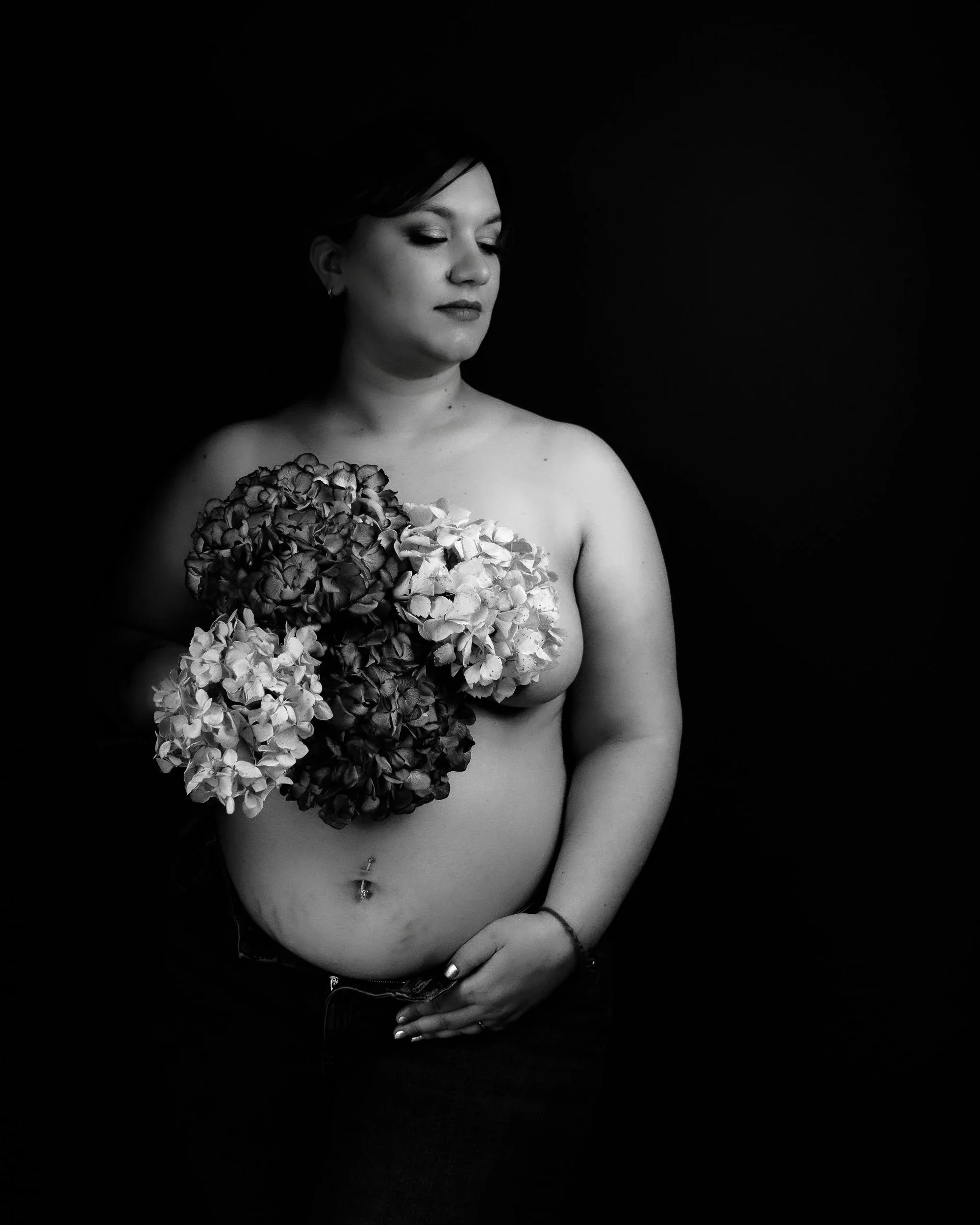 Une femme nue tenant un bouquet de fleurs, photographiée en noir et blanc contre un fond noir.
