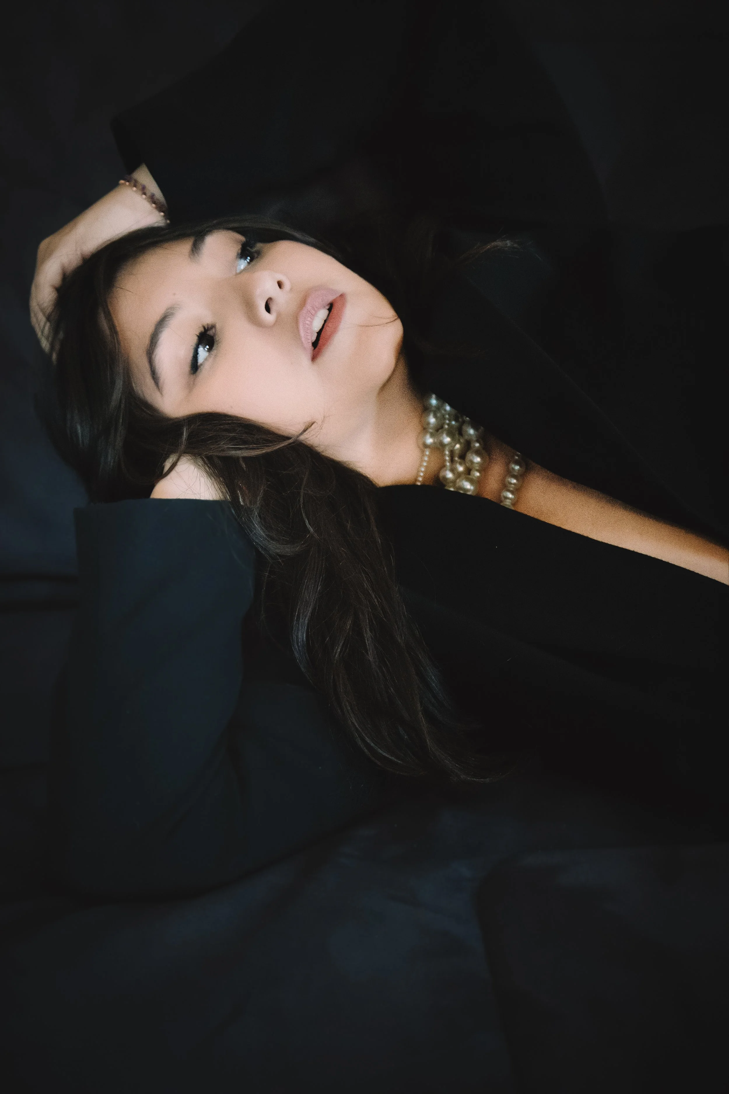 Une femme élégante avec des perles, portant un vêtement noir, avec des cheveux bruns longueur moyenne, allongée sur un fond sombre.