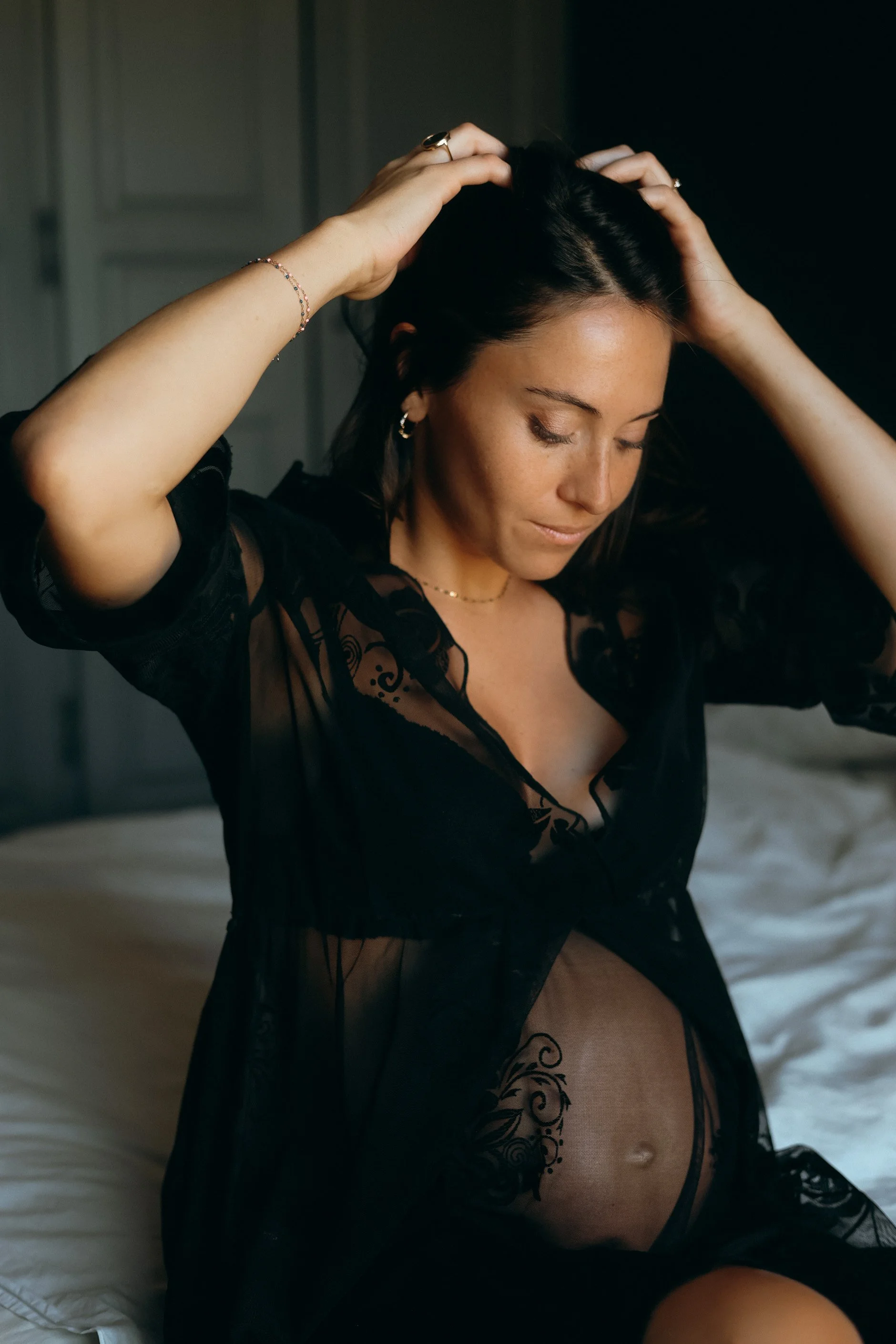 Une femme en lingerie noire transparente, assise sur un lit, les mains dans les cheveux, regardant vers le sol, dans une pièce sombre.