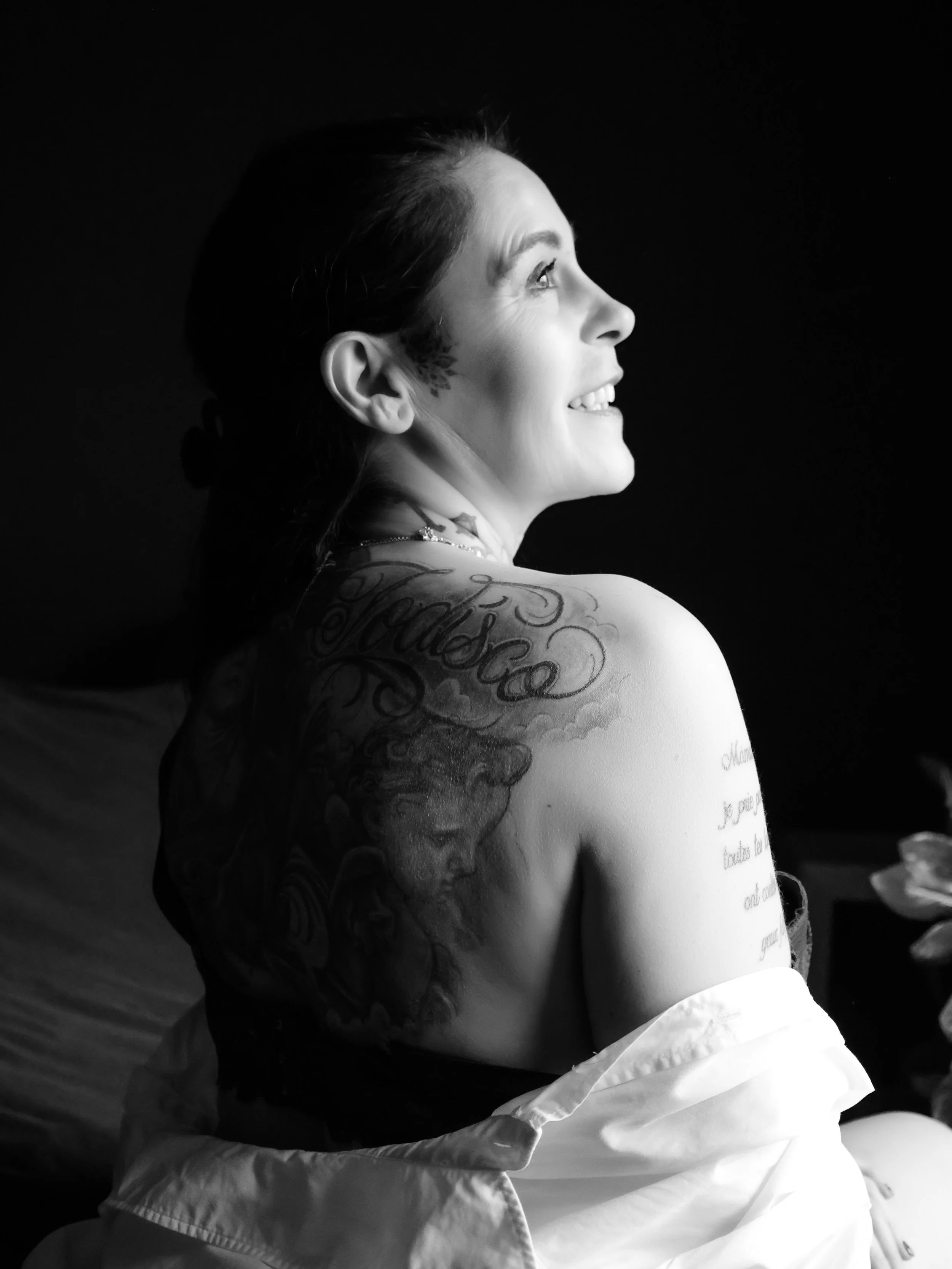 Femme souriante avec tatouages sur le bras et l'épaule, vue de profil en noir et blanc, assise avec une chemise ouverte.