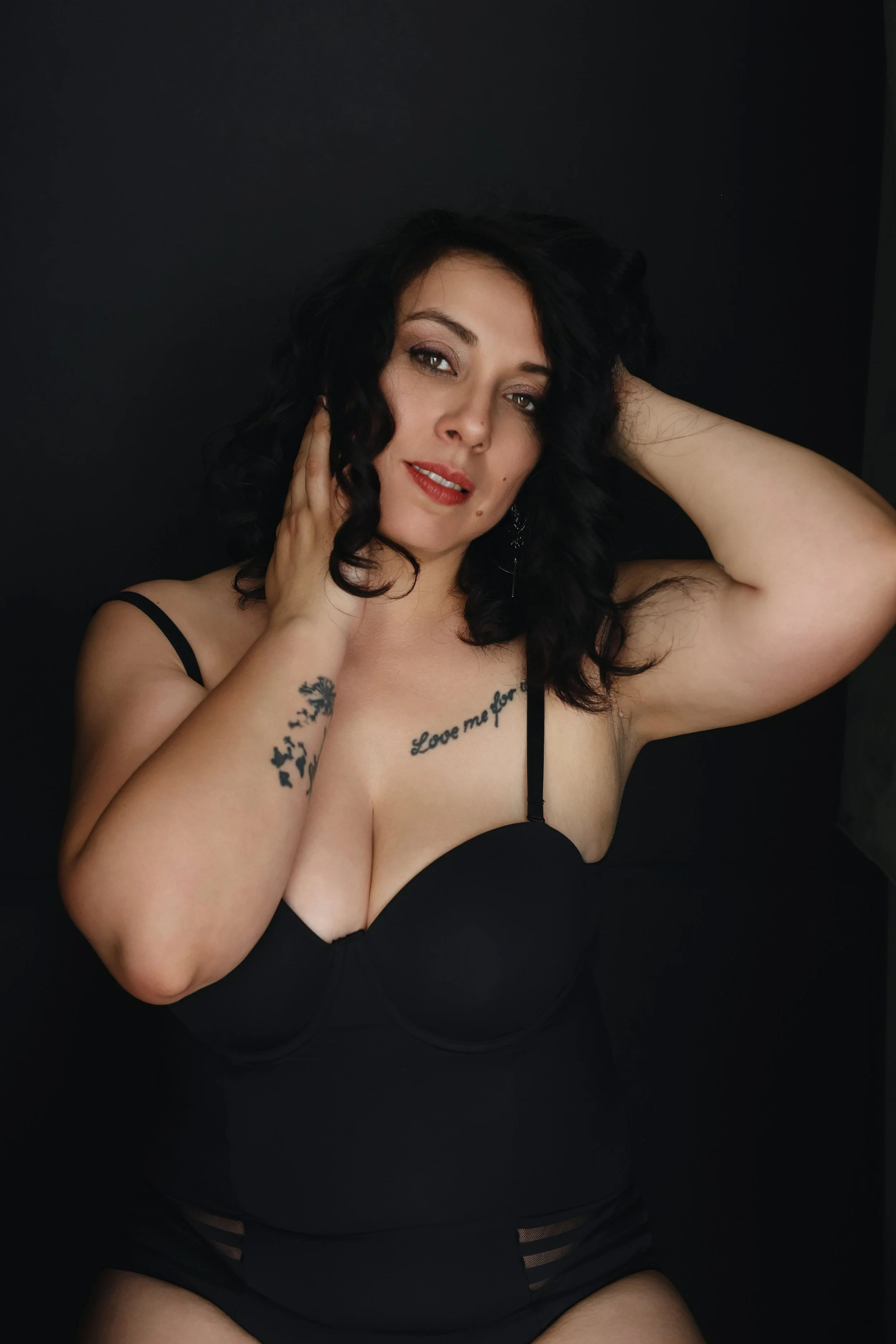 Femme aux cheveux noirs bouclés, portant une tenue noire, posant contre un fond noir, tenant sa tête d'une main et touchant son visage de l'autre.