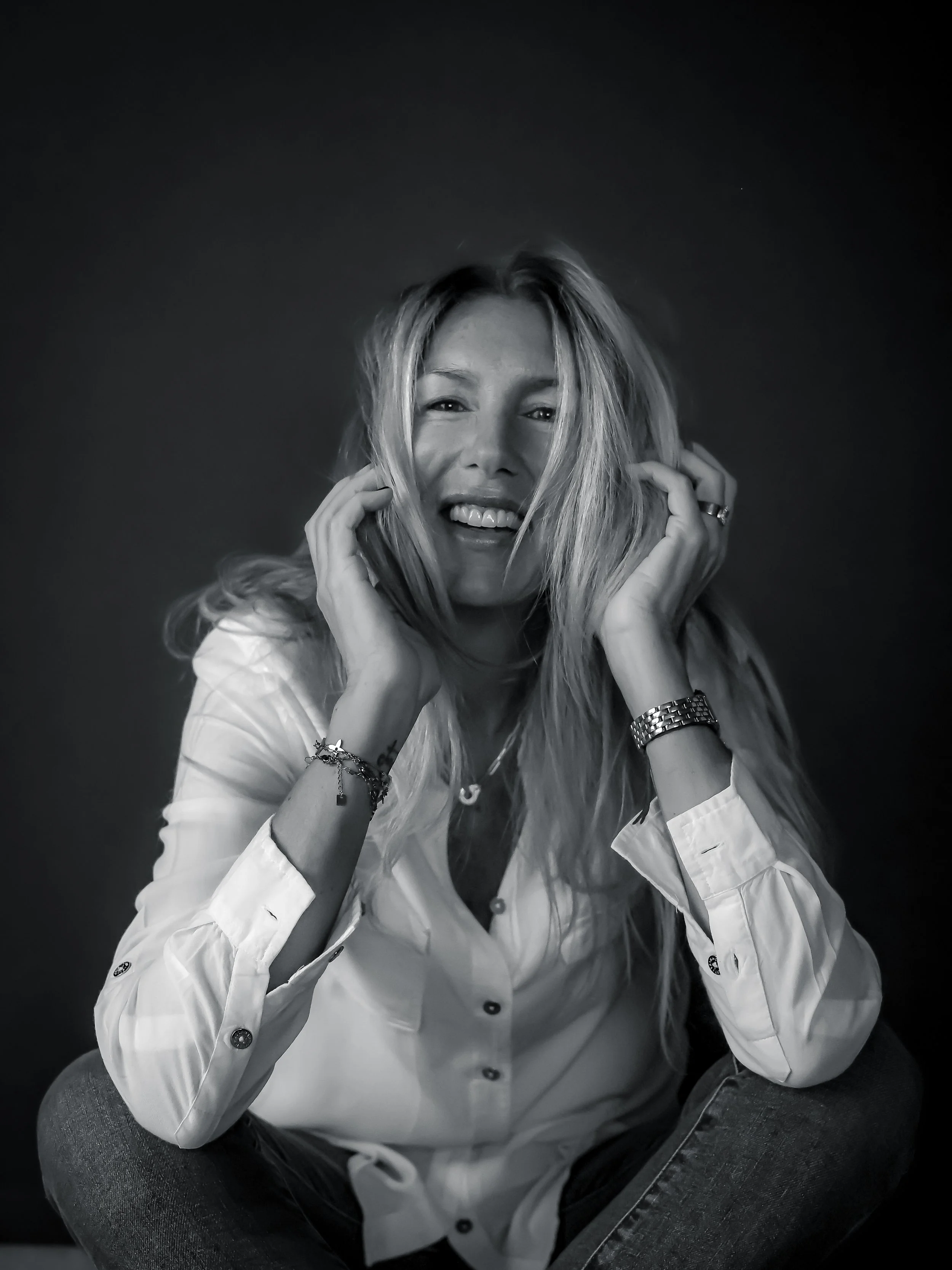 Femme souriante avec cheveux longs, portant une chemise blanche, chaussant un jean, assise en posture décontractée contre un fond sombre.