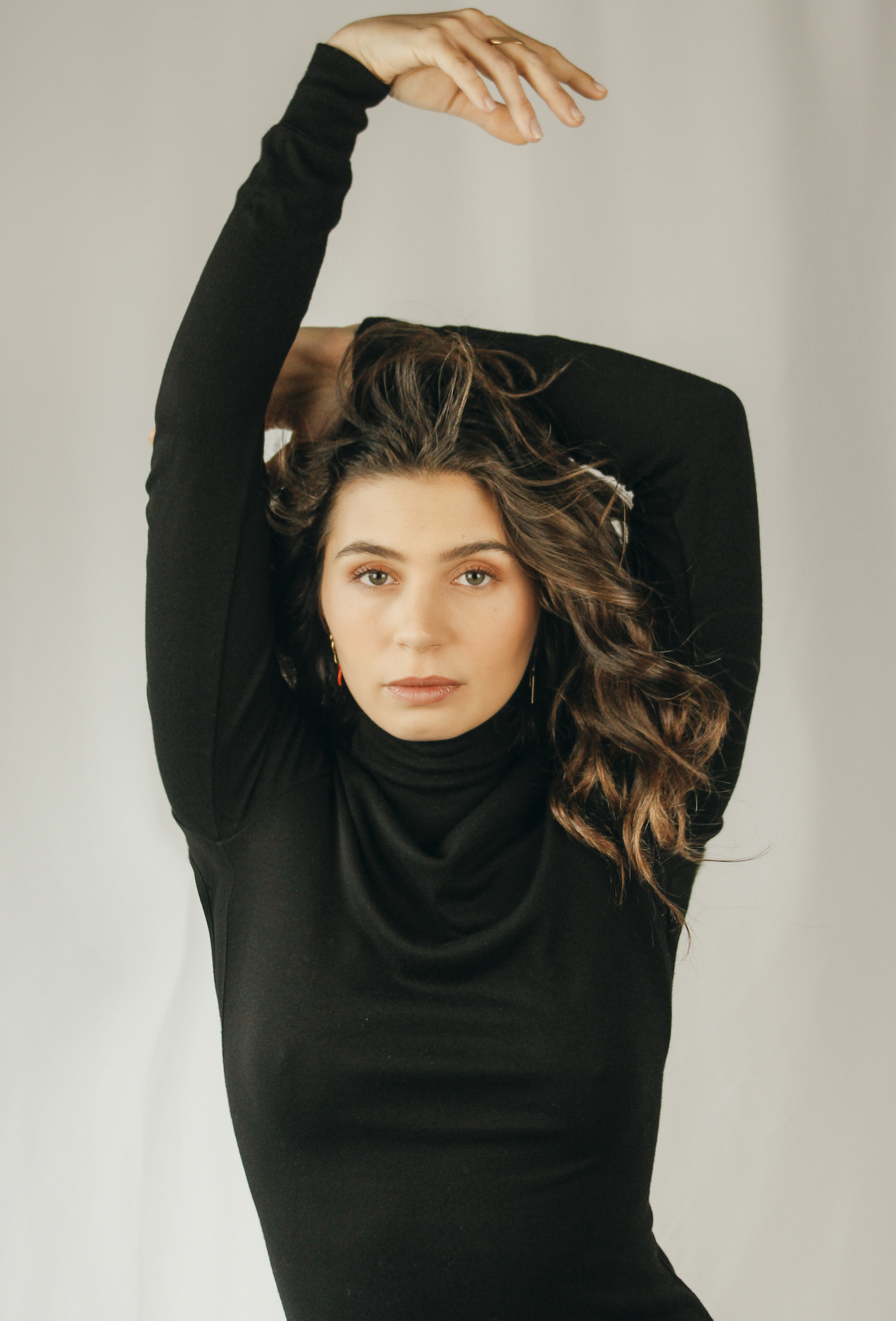 Une femme avec des cheveux bruns bouclés porte un haut noir et regarde directement l'objectif, levant un bras au-dessus de sa tête dans une pose élégante.