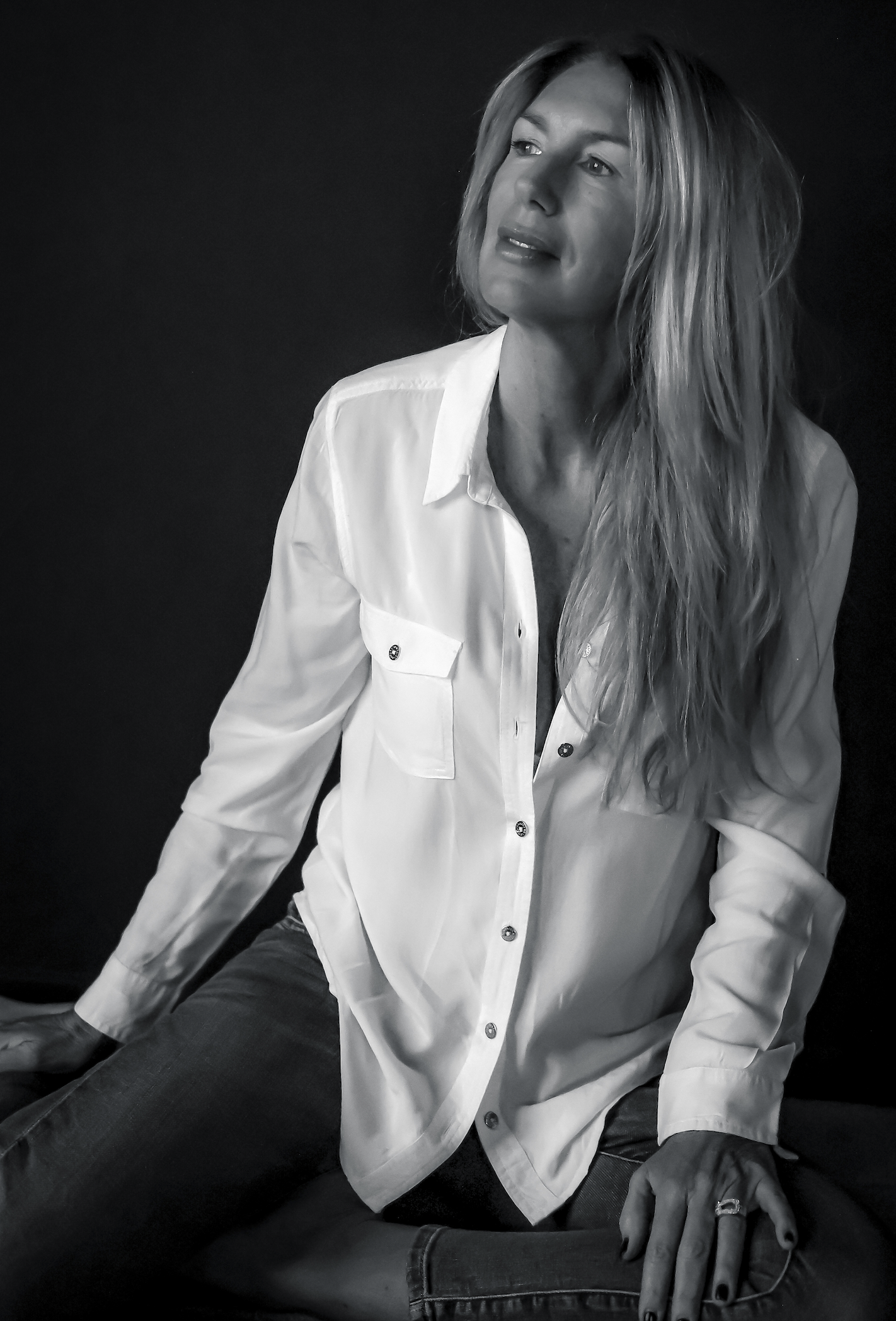 Femme assise portant une chemise blanche, cheveux longs et ondulés, regardant vers la gauche, fond noir.