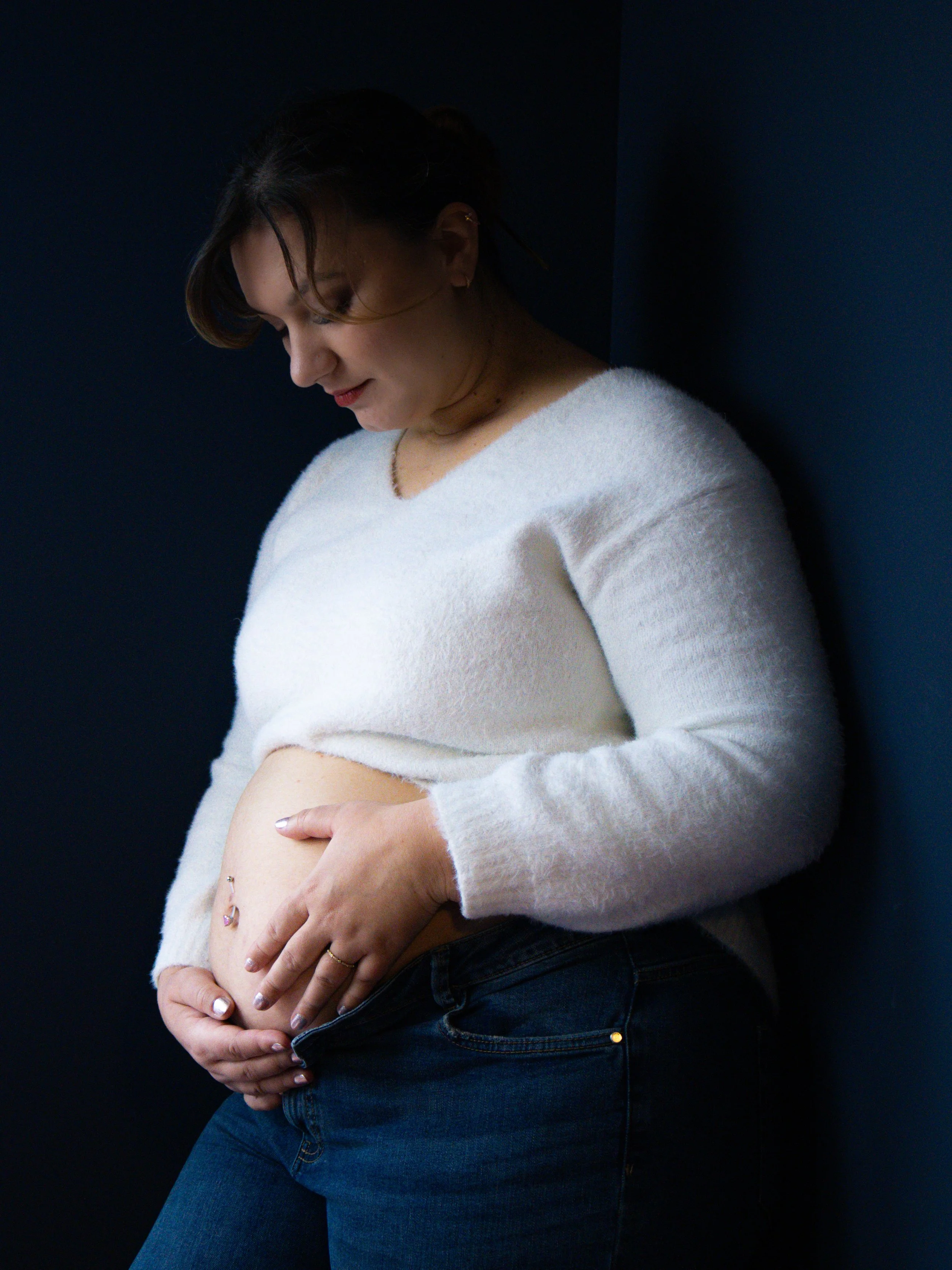 Femme souriante portant un pull blanc, mettant sa main sur son ventre arrondi, debout contre un mur sombre.