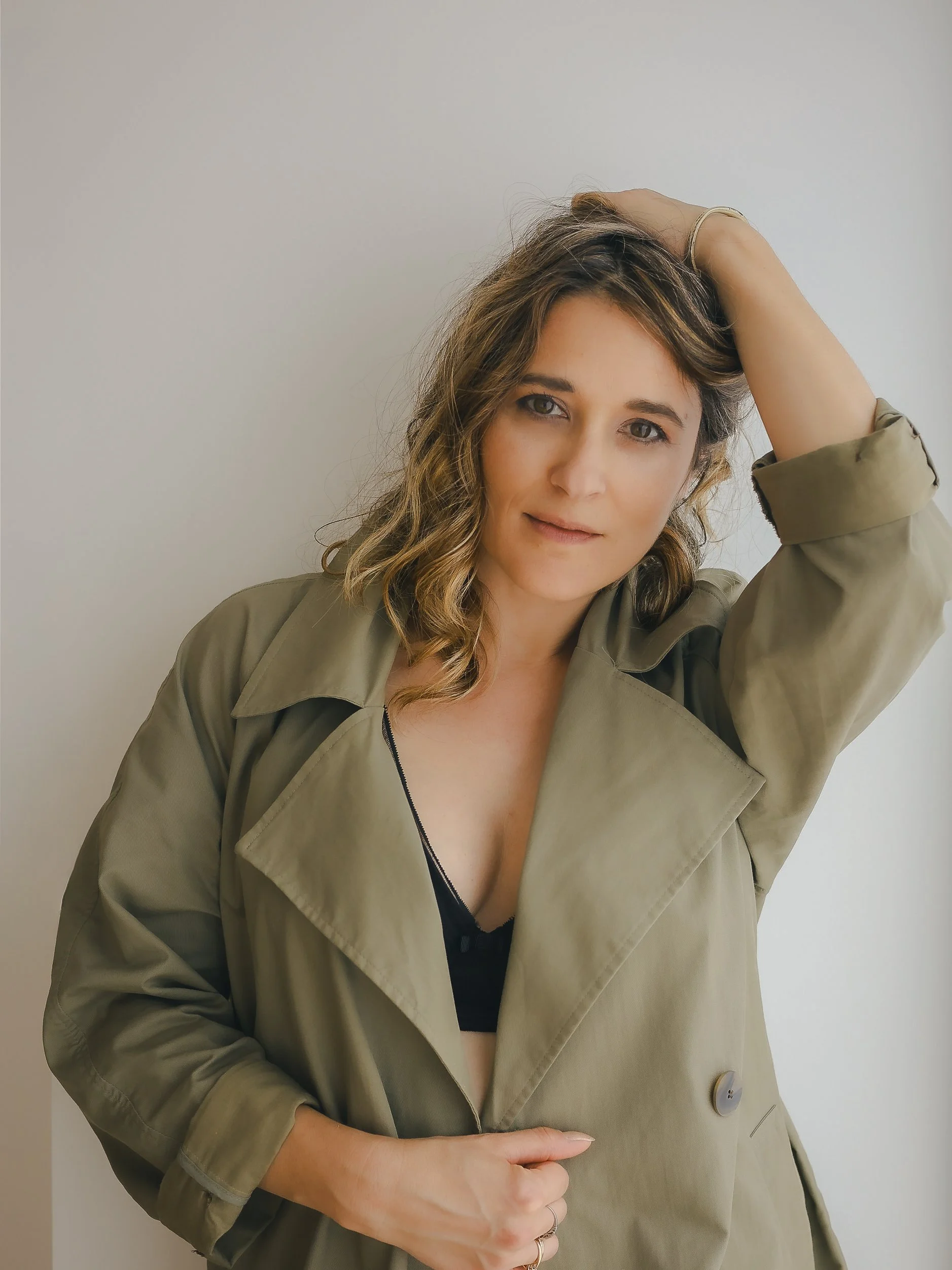 Jeune femme avec des cheveux bouclés, portant una veste beige et une lingerie noire, regardant l'objectif avec une expression neutre, contre un fond blanc.