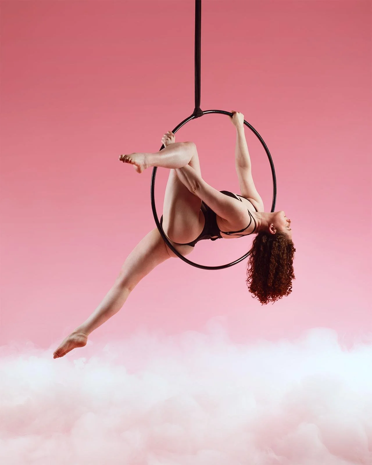 A woman with curly hair performing an aerial circus act on a black hoop suspended in the air with a pink background and clouds at the bottom.