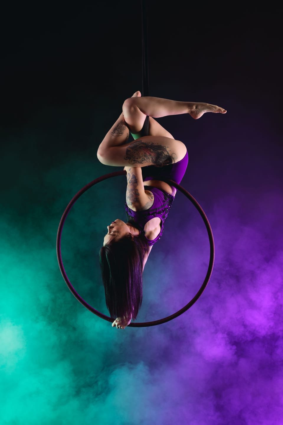 A woman performing an aerial hoop act with colorful stage lighting and mist in the background.