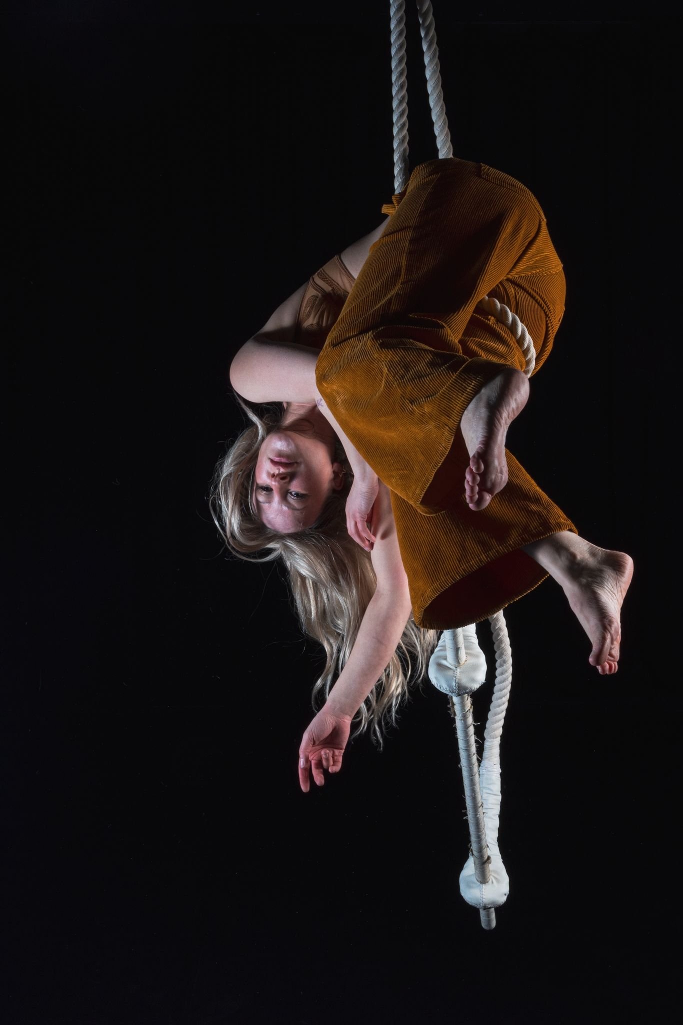 A woman with long blonde hair hanging upside down from a trapeze against a black background.