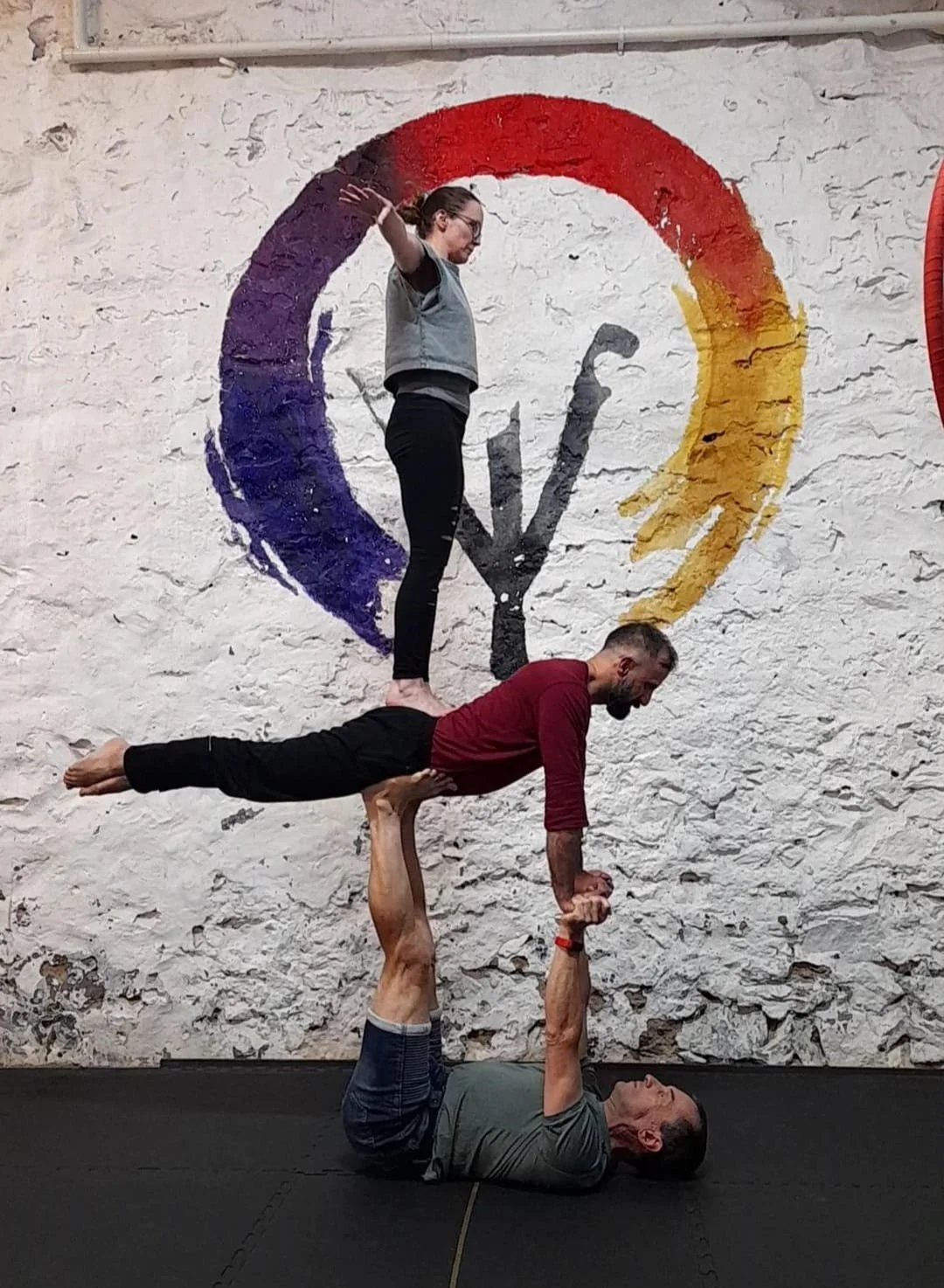 Four people are performing an acro yoga pose indoors against a white brick wall with a colorful circular logo painted on it. One person is lying on the floor on their back, supporting another person on their hands and feet in a horizontal plank position. A third person is balancing on the second person's hands, standing with arms extended to the sides.