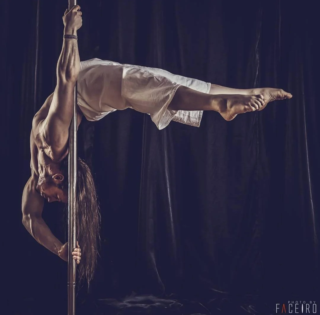 A man performing a pole dance move, hanging upside down with his legs extended horizontally and holding the pole with his hands, wearing white shorts and a sleeveless top, against a dark background.