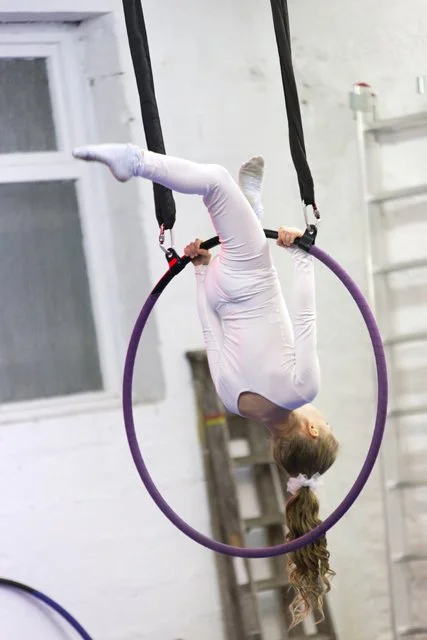 A young girl in a white gymnastic outfit practicing on an aerial hoop in a gym.