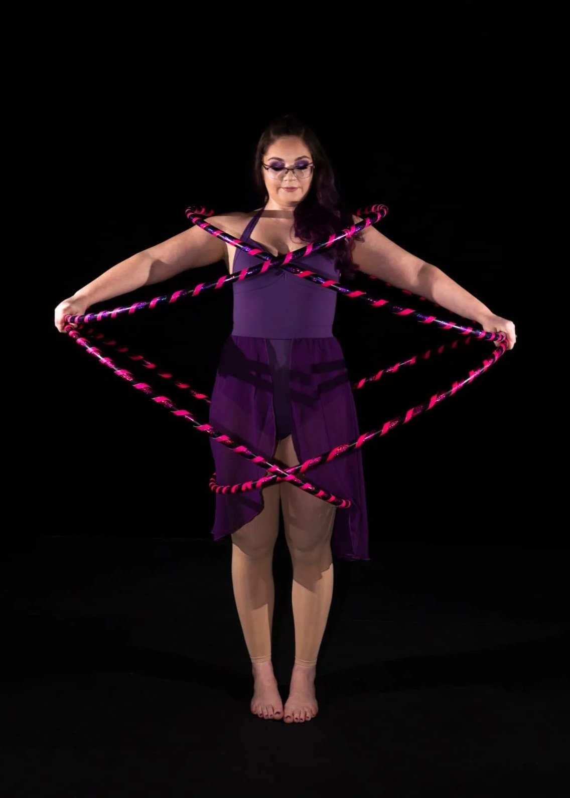 A woman stands on a black background, holding four pink and black striped hula hoops around her body.
