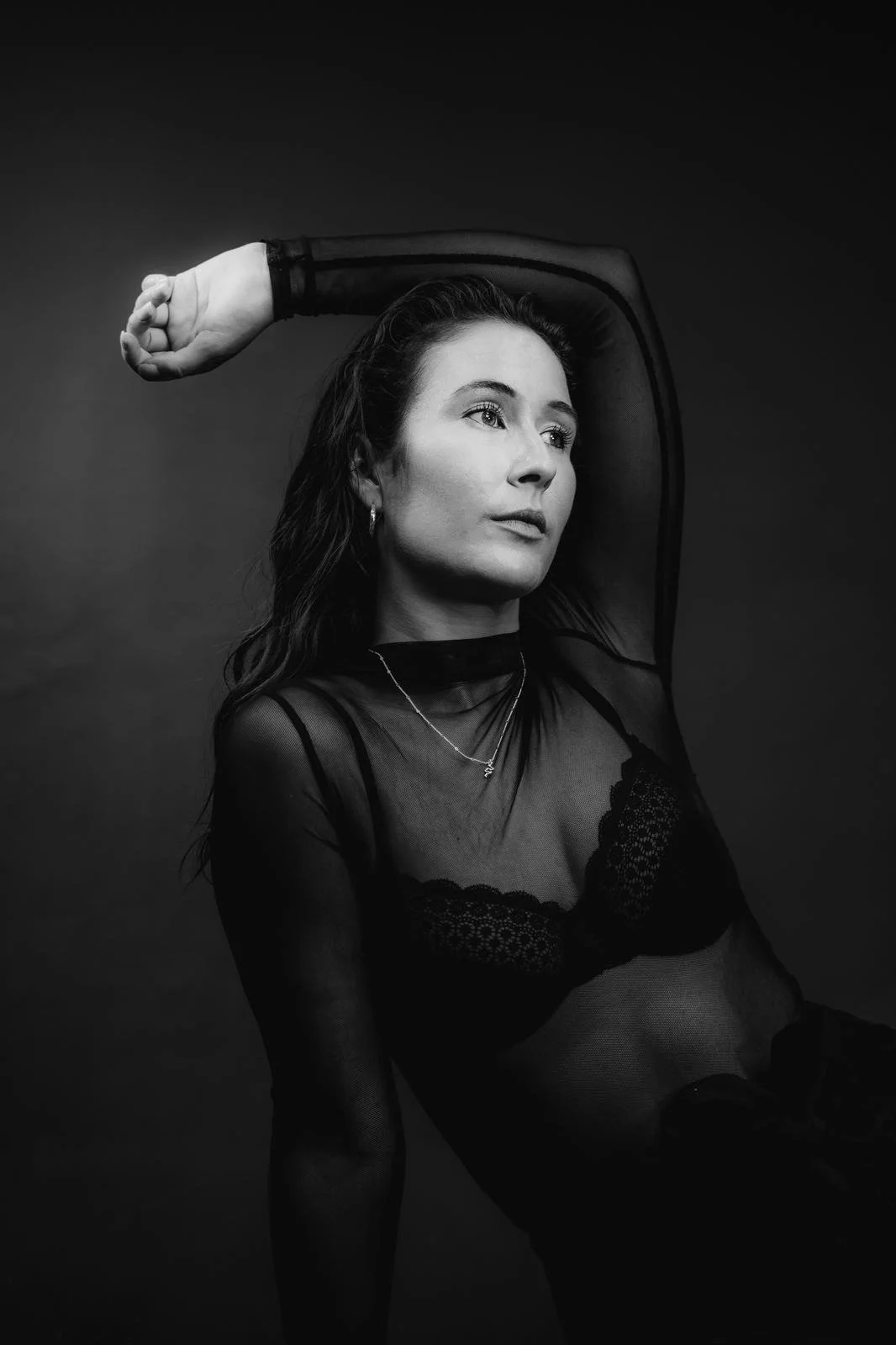 A black and white portrait of a woman with wavy hair, wearing a sheer top and leaning back with her arm raised above her head.
