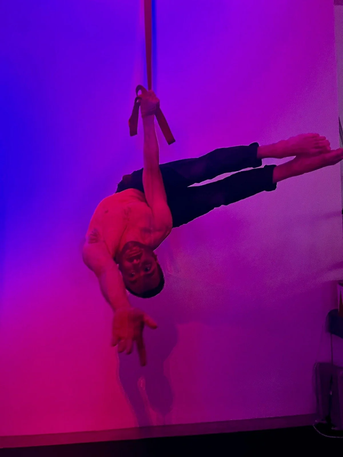 A man performing a side plank yoga pose, hanging from a strap attached to the ceiling, in a dimly lit room with purple and pink lighting.
